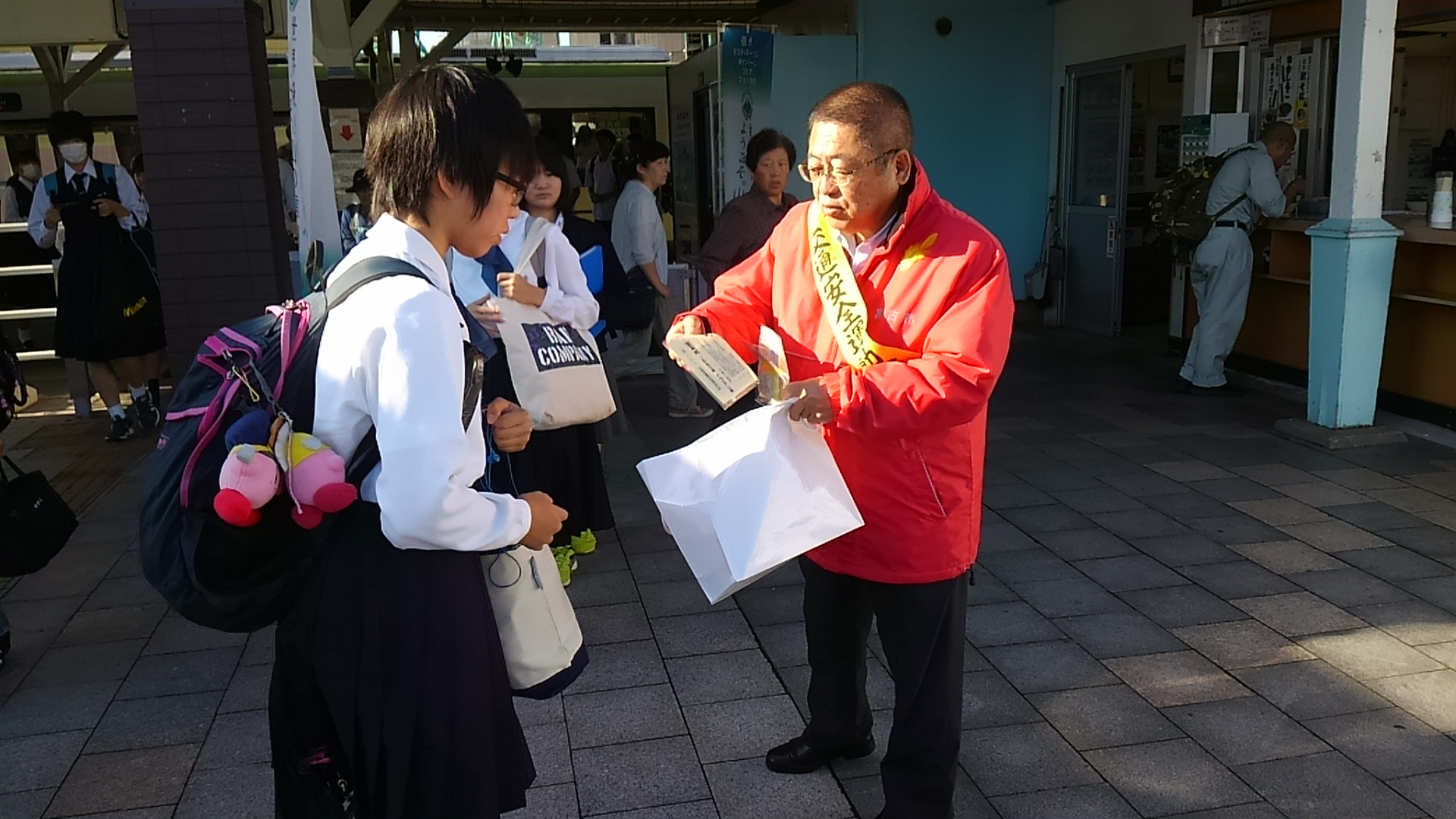 オレンジ色のジャンパーに黄色のタスキをかけた市長が、白い紙袋の中から出した反射シールを女子学生に配布している様子の写真