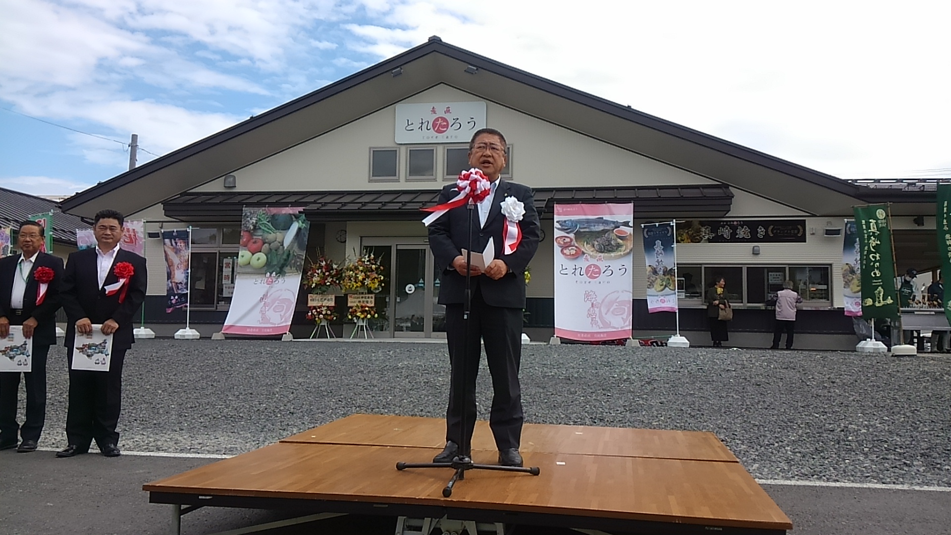 三角屋根の下に「産直とれたろう」と書かれた看板が設置された建物をバックに、マイクスタンドが設置された台の上に立つ市長が挨拶を行っている様子の写真