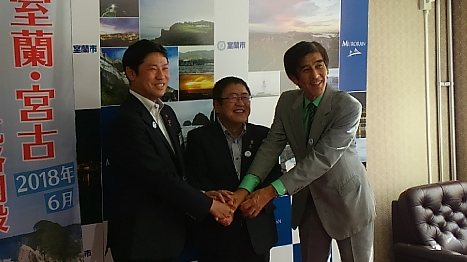 室蘭市内の風景の写真が掲載されたポスターの前で、市長を中央に室蘭市の青山市長と栗林商工会議所会頭が笑顔で手を取り合っている記念写真