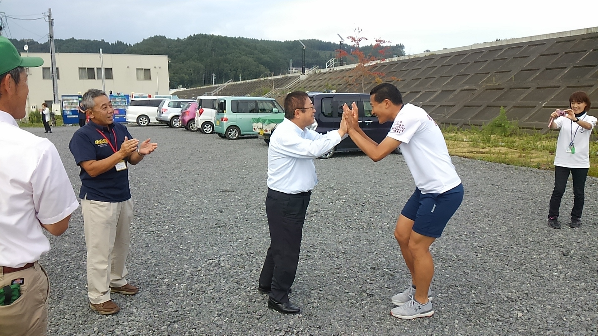 関係者の男性が拍手をするなか、白いシャツに紺色の短パンを履いた男性と市長がハイタッチをしている様子を右側の女性が撮影している写真