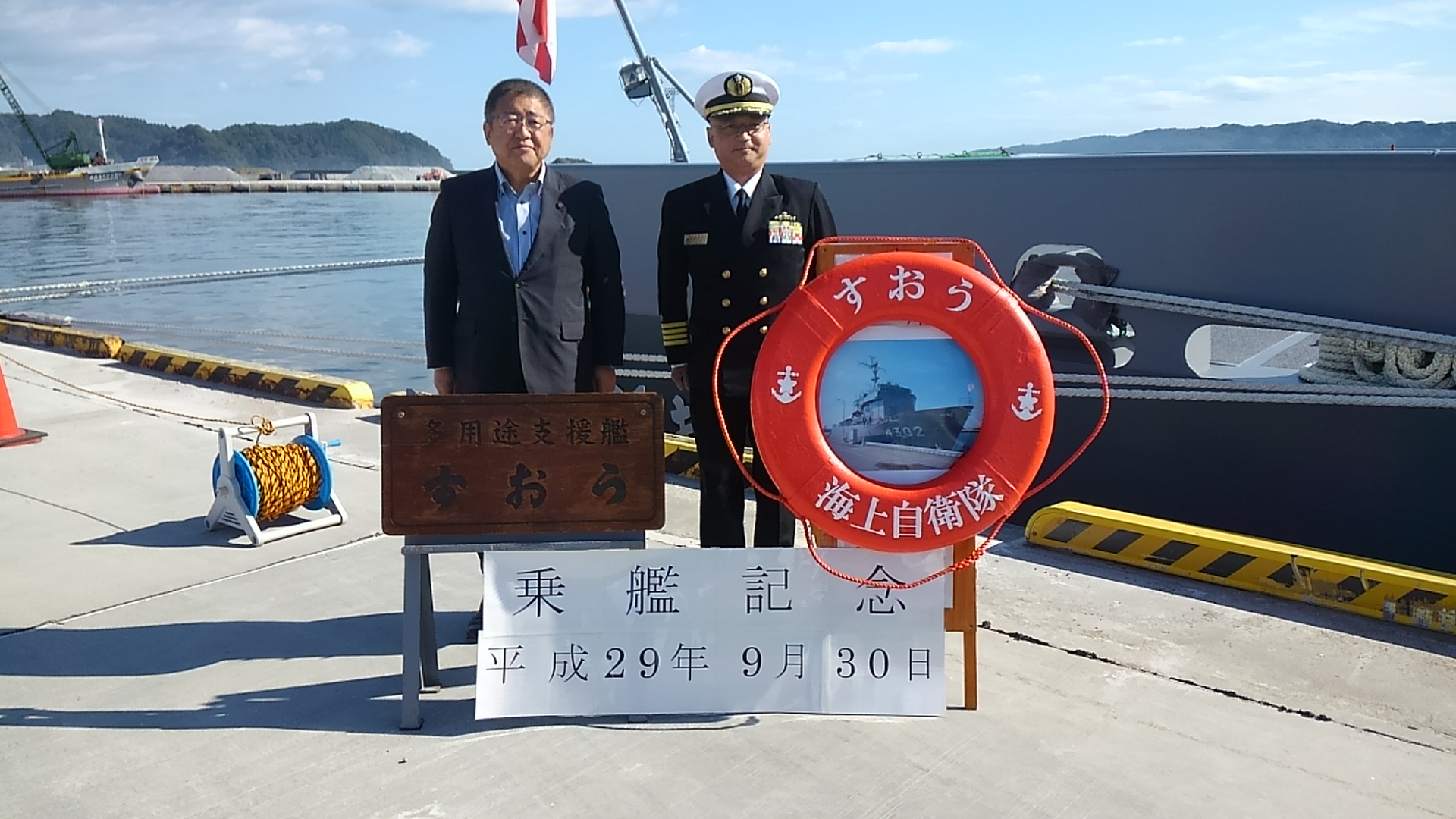 藤原埠頭に入港した海上自衛隊多用途支援艦「すおう」をバックに、碇のマークと「すおう海上自衛隊」と書かれたオレンジ色の浮き輪の様なものと、「乗艦記念 平成29年9月30日」と書かれた横幕が貼られた後ろに大湊警備隊司令加藤一等海佐と市長が並んで立っている写真
