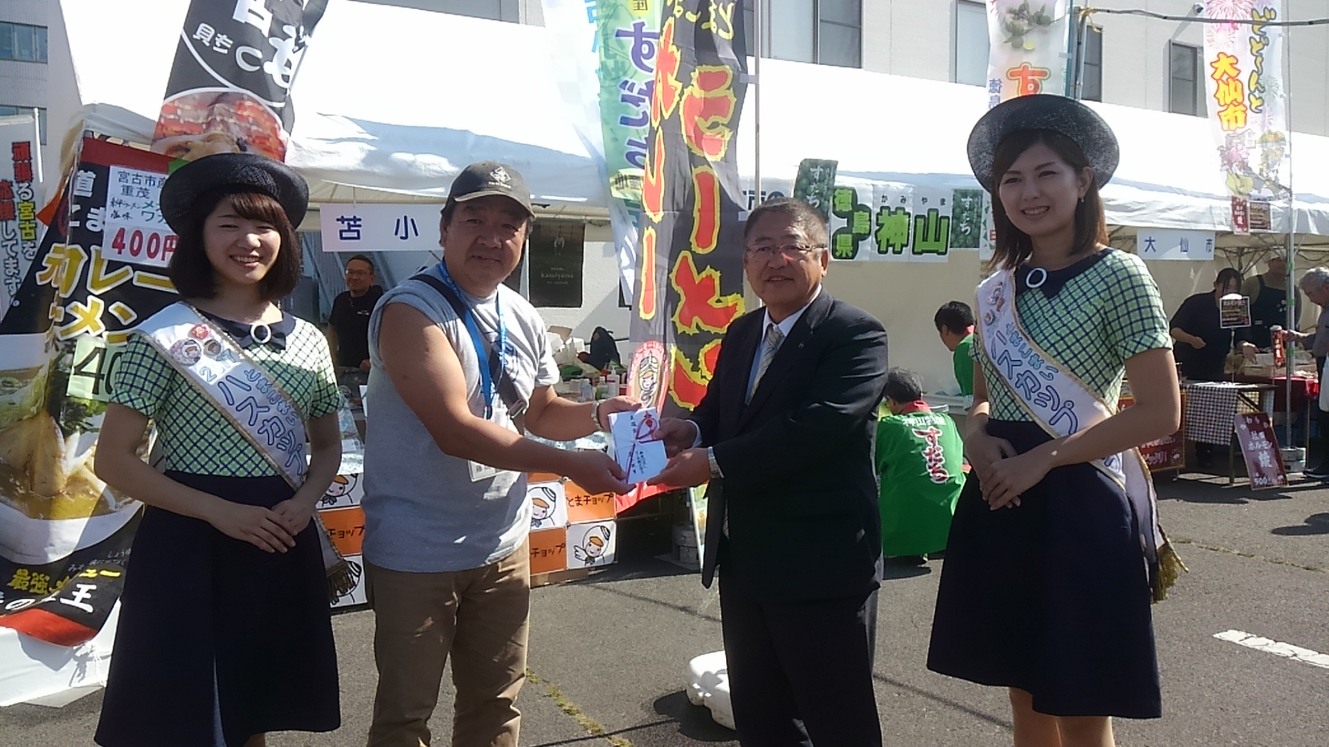苫小牧市の販売ブースの前で、2名のハスカップレディの間に黒色のキャップを被った苫小牧観光協会の男性と市長が頂いた支援金を一緒に持っている記念写真
