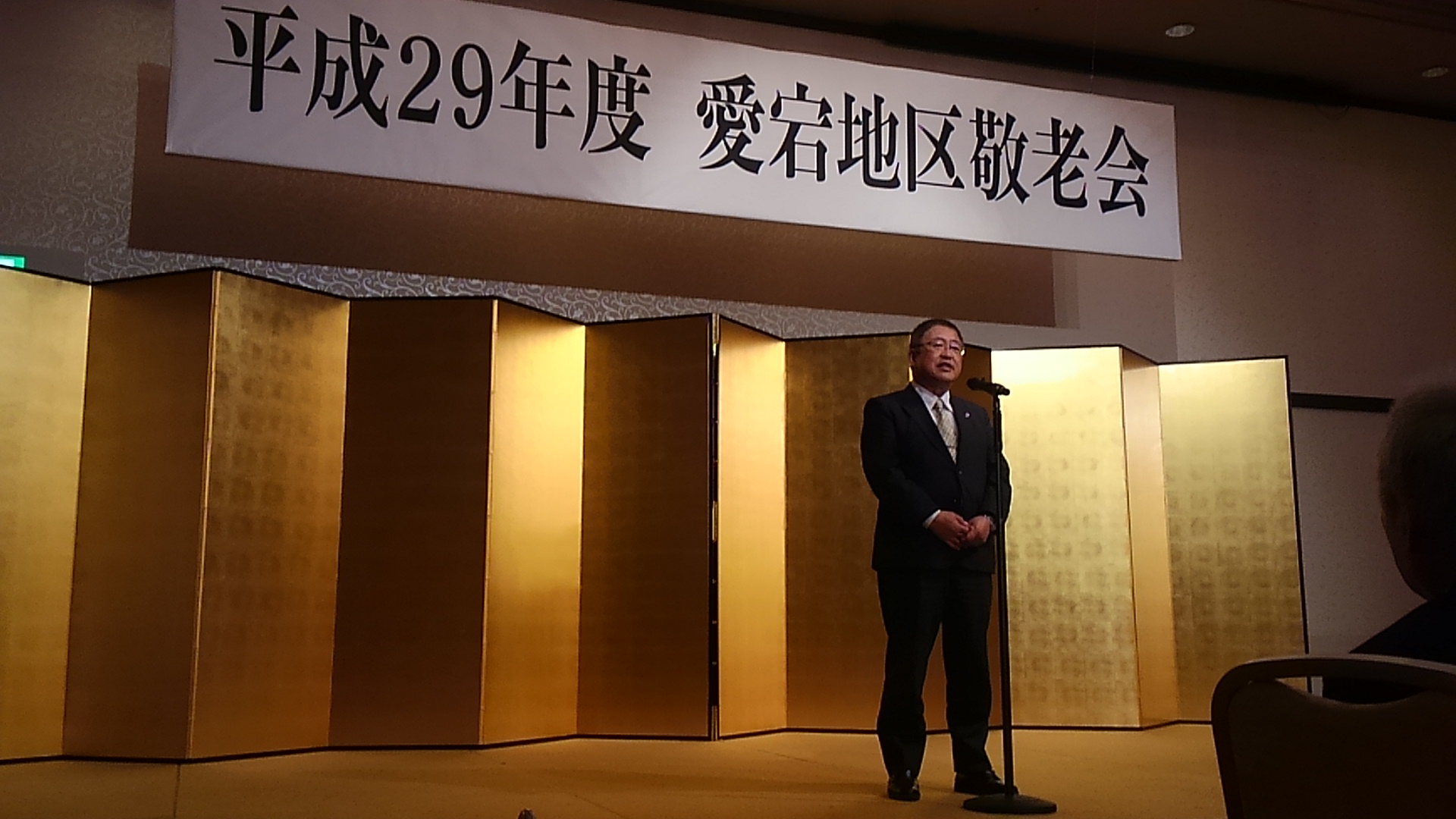 「平成29年度 愛宕地区敬老会」と書かれた横断幕と金屏風が置かれた舞台上で、スタンドマイクに向かい話をする市長の写真