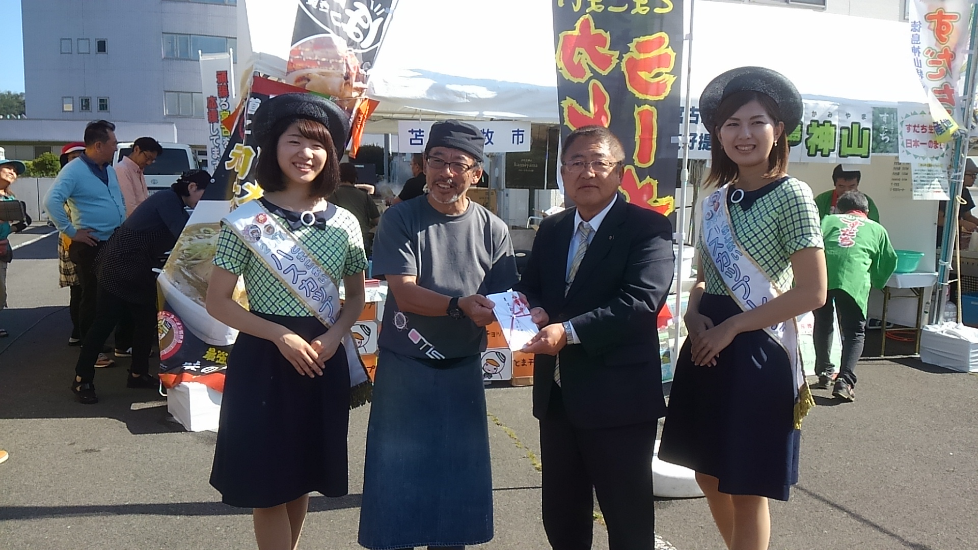 苫小牧市の販売ブースの前で、2名のハスカップレディの間に頭に黒色のバンダナを巻いたとまこまい味な倶楽部の男性と市長が頂いた支援金を一緒に持っている記念写真