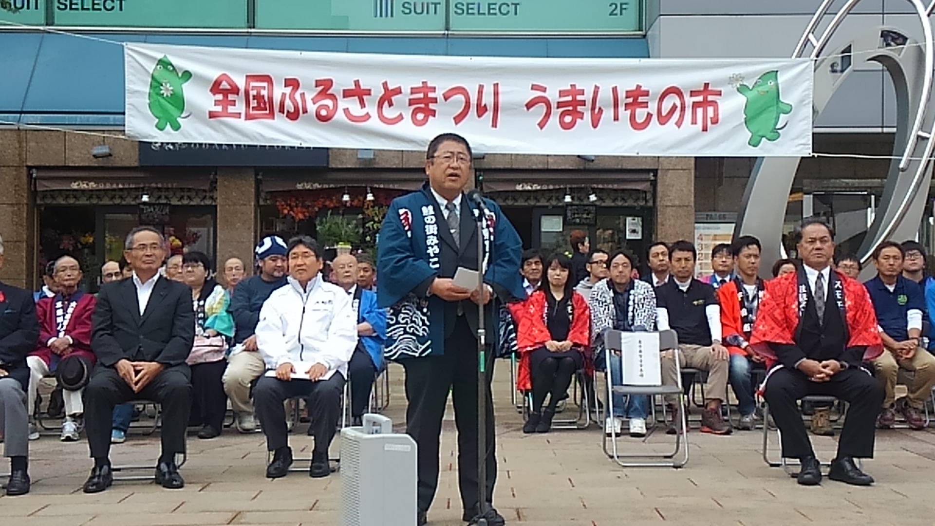 「全国ふるさとまつり うまいもの市」という横断幕の前で椅子に座る参加者の方々と、スタンドマイクに向かい話をする市長の写真