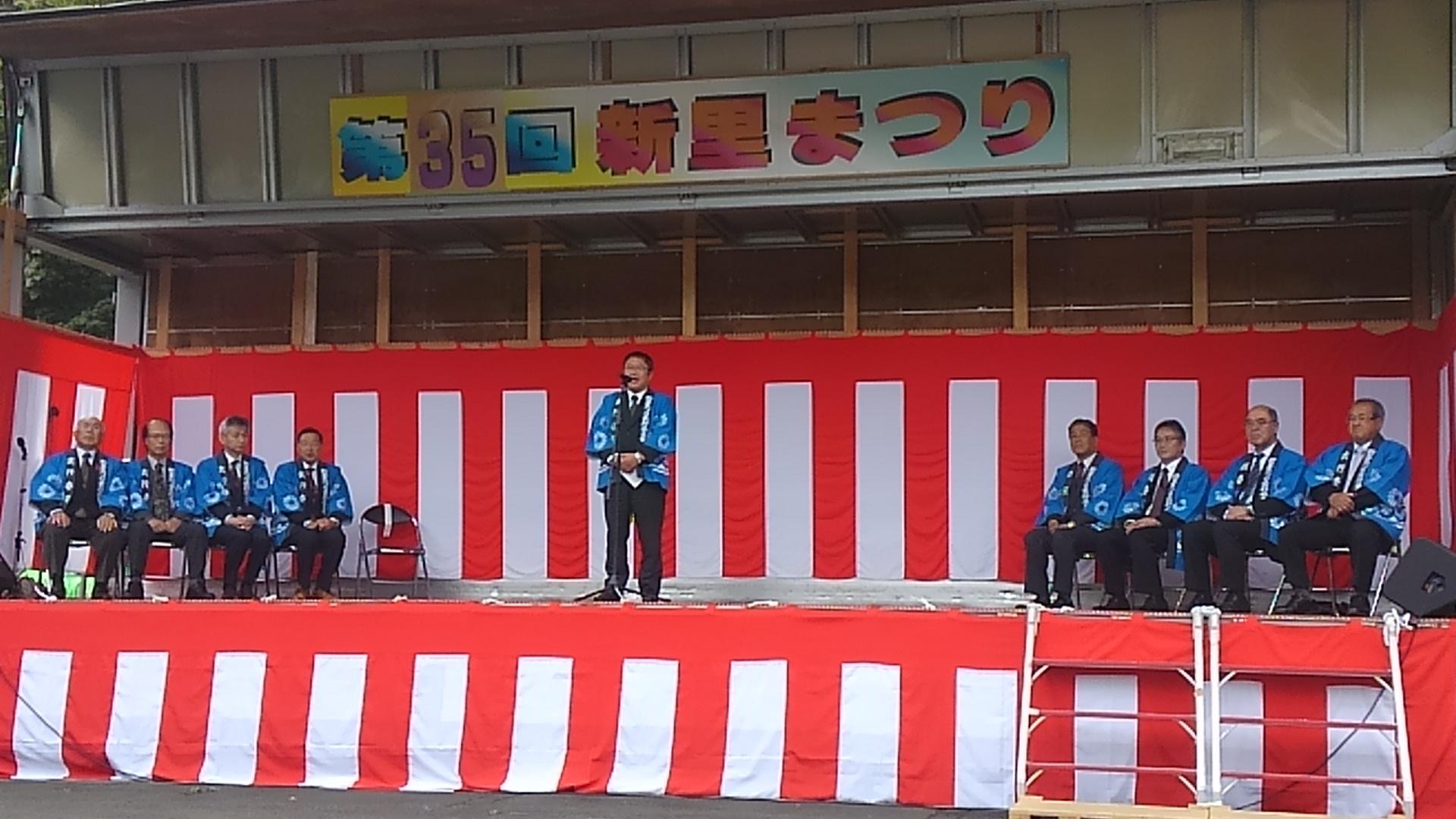 「第35回 新里まつり」と書かれた看板の下で、紅白幕の貼られた舞台の左右に4名ずつ座る青い法被を着た男性と、中央でスタンドマイクに向かい話をする青い法被を着た市長の写真