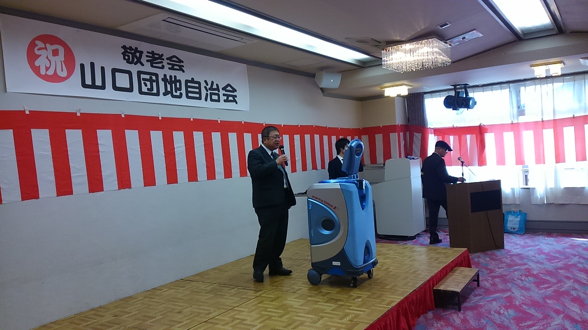 舞台の上で、カラオケの機械の前に立ち歌う様子の市長の写真