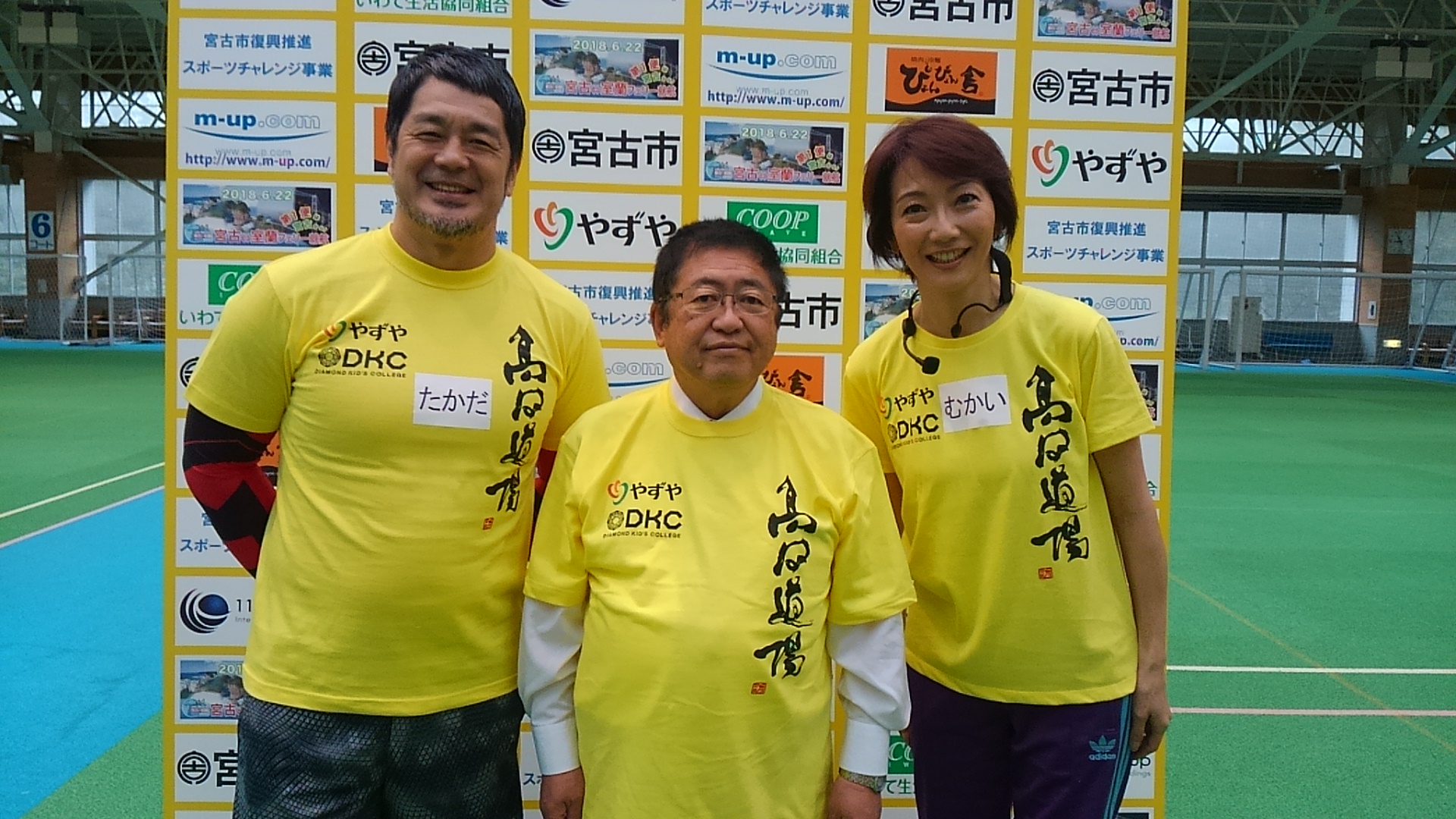 お揃いの黄色のTシャツを着てバックパネルの前で並ぶ市長、高田延彦さん、向井亜紀さんの記念写真