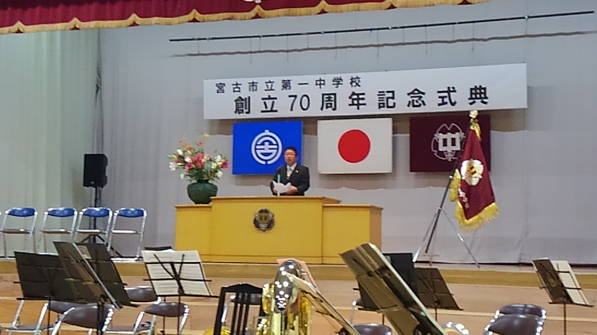 「宮古市立第一中学校 創立70周年記念式典」と書かれた横断幕や旗が掲げられた舞台上で、演台のマイクに向かい話をする様子の市長の写真