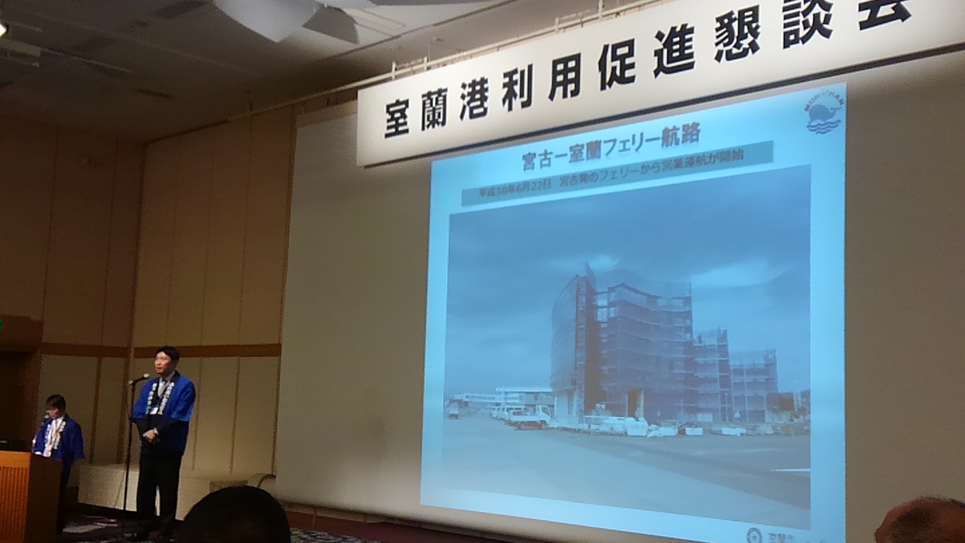 「室蘭港利用促進懇談会」と書かれた横断幕と画面が映されたスクリーンの横で、青い法被を着てスタンドマイクに向かい話をする男性の写真