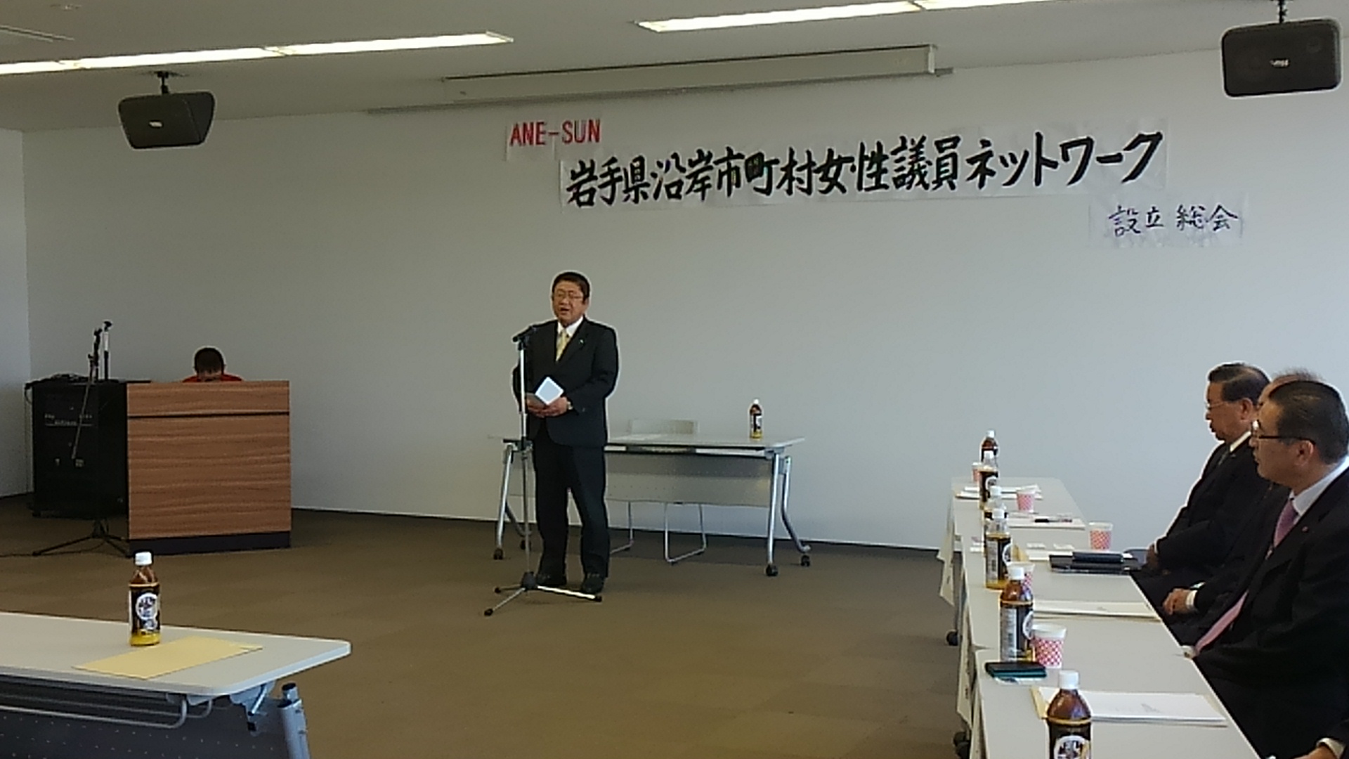 「岩手県沿岸市町村女性議員ネットワーク設立総会」と書かれた紙が貼られた壁の前で、スタンドマイクに向かい話をする市長の写真
