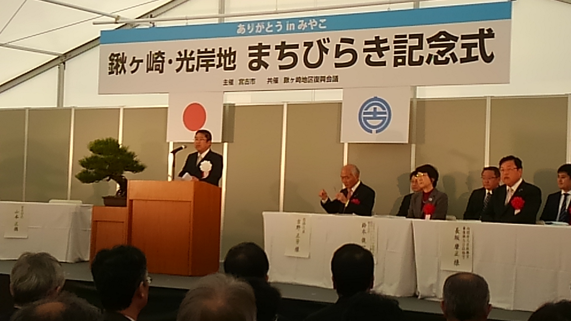 「鍬ヶ崎・光岸地まちびらき記念式」と書かれた横断幕と日本国旗、宮古市の市町村旗が掲げられた会場で、白いテーブルクロスが敷かれた机の席に座る関係者の方々と、演台のマイクに向かい話をする様子の市長の写真