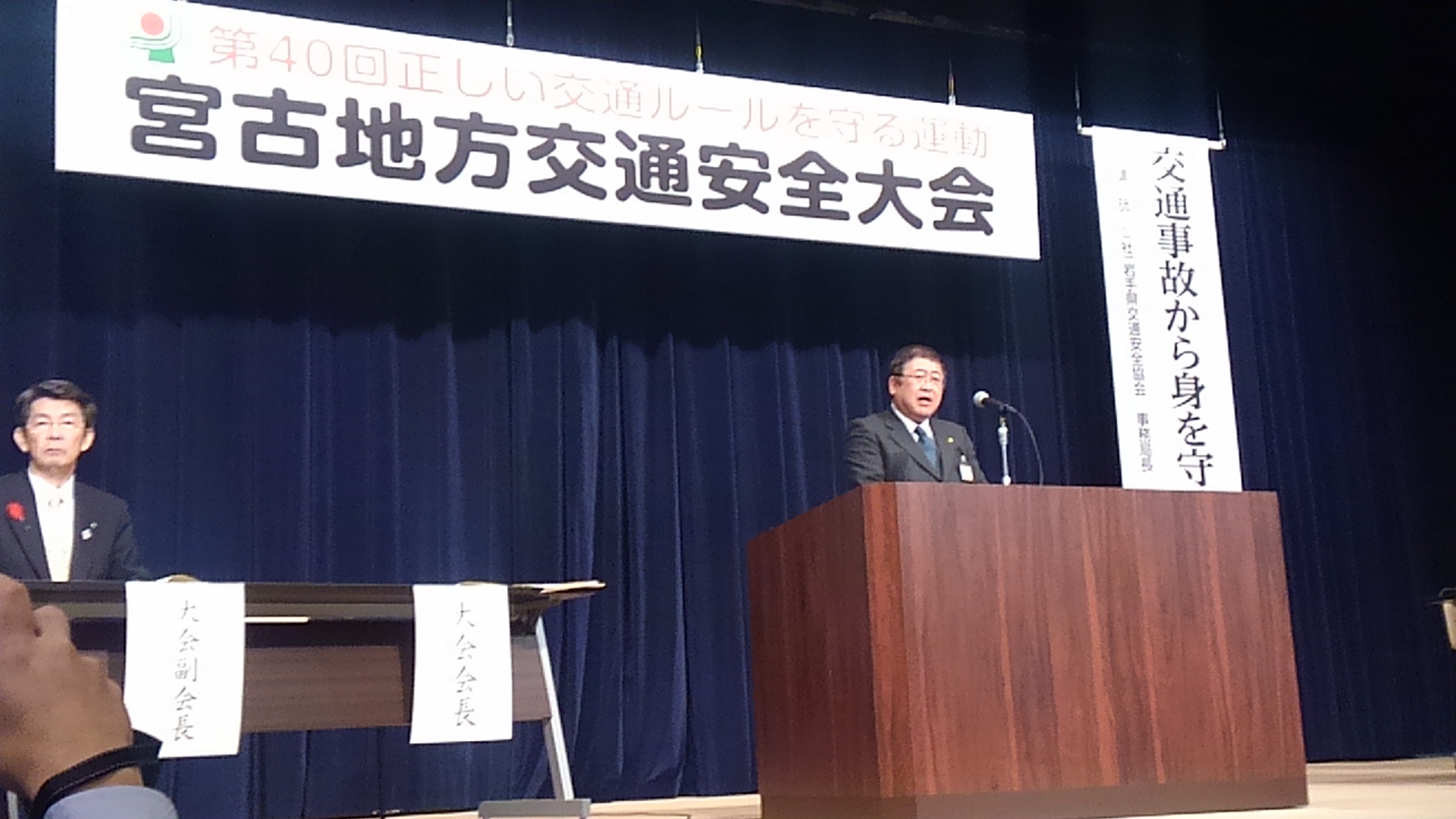 「宮古地区交通安全大会」と書かれたパネルが吊るされた舞台上で、演台に置かれたマイクに向かい話をする様子の市長の写真