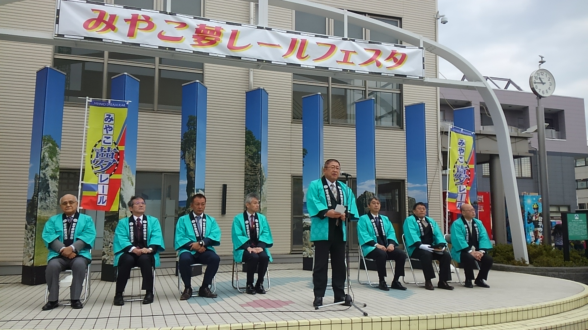 「みやこ夢レールフェスタ」と書かれた横断幕の下で、青い法被を着て椅子に座る7名の男性と、スタンドマイクに向かい話をする市長の写真