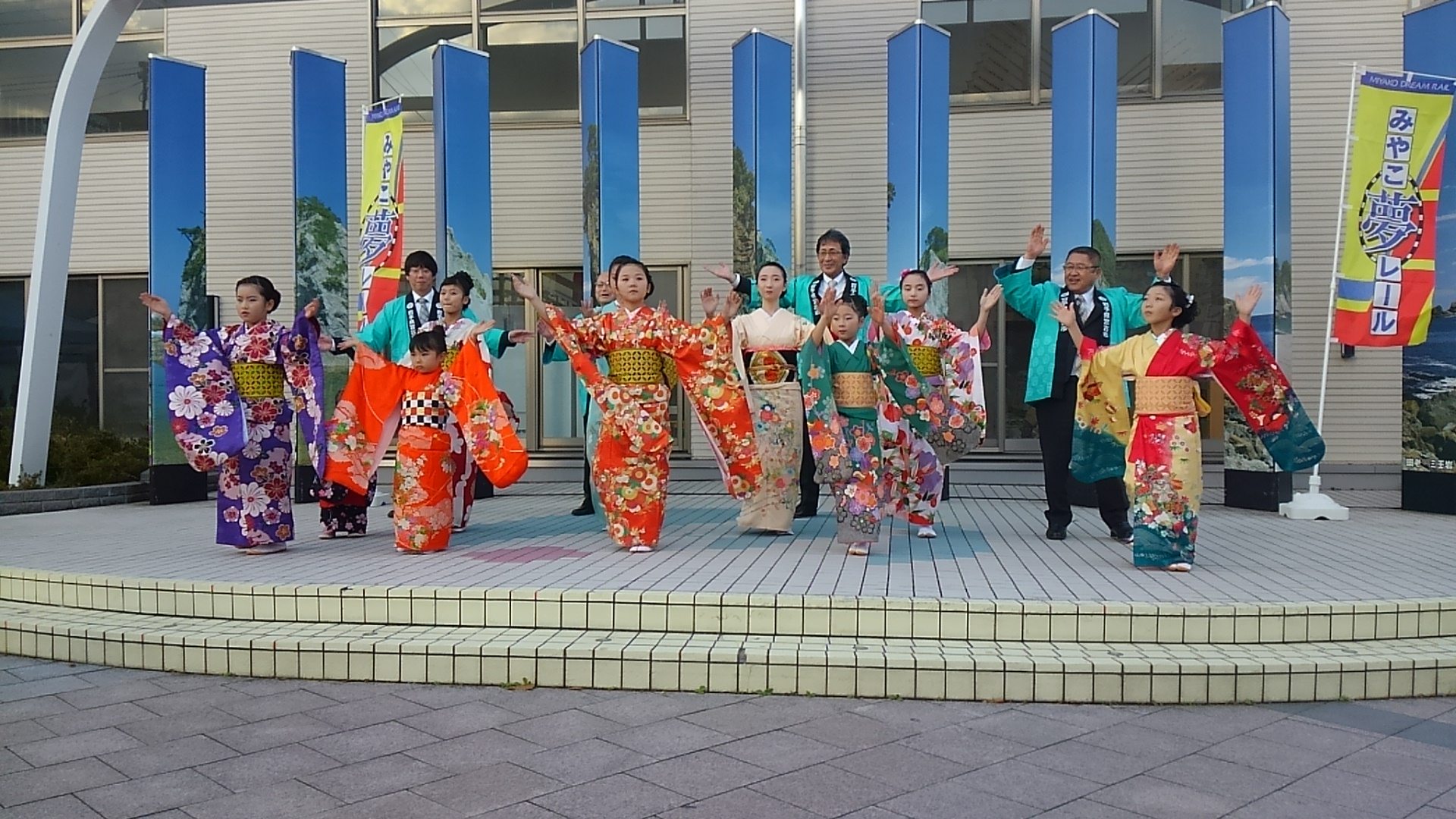 着物を着て踊る女の子たちと、後方で踊る市長と関係者の男性3名の写真