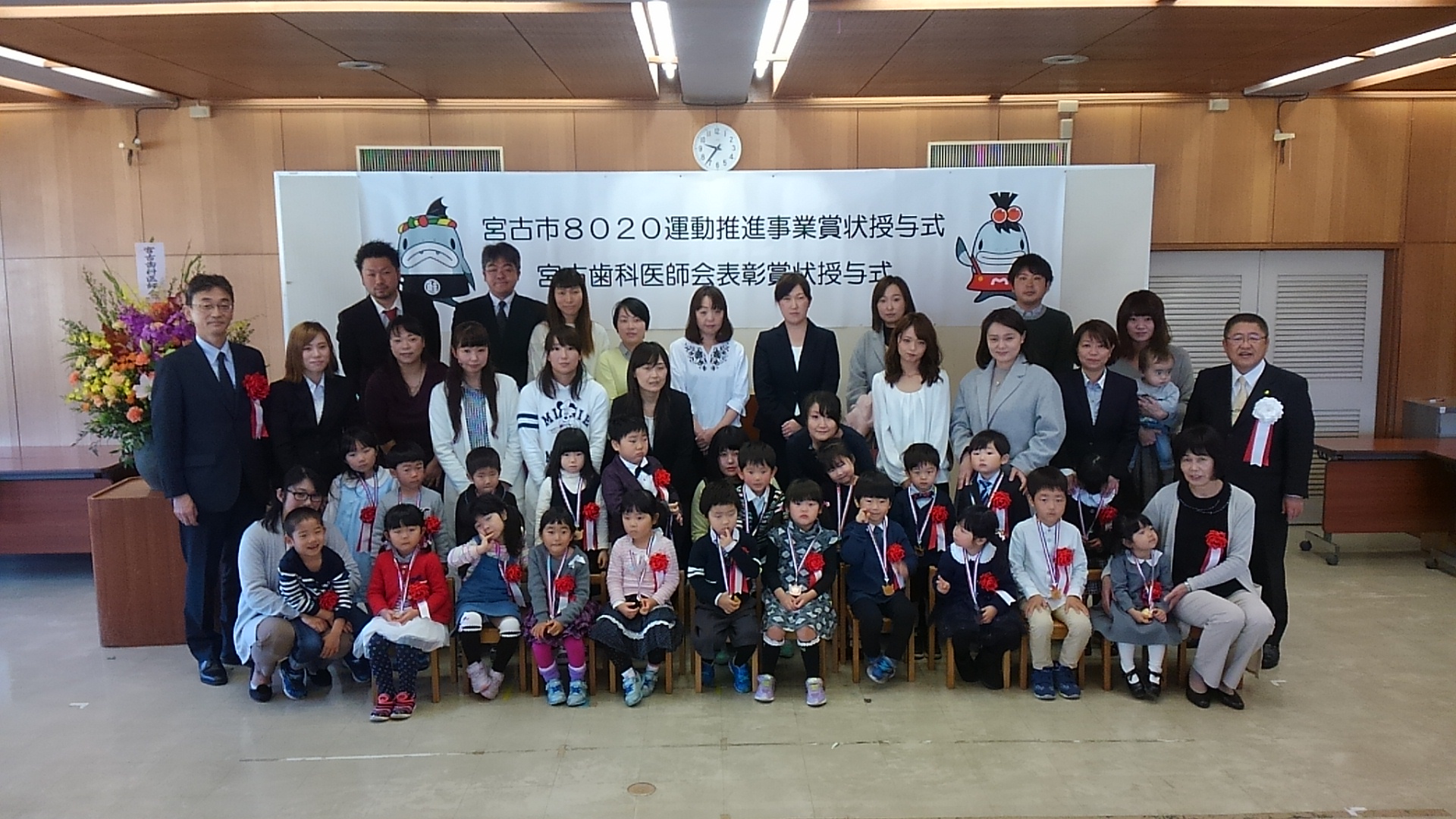 胸章を着けて首からメダルを下げた大勢の子供たちと、保護者の女性や男性たちが並んでいる記念写真