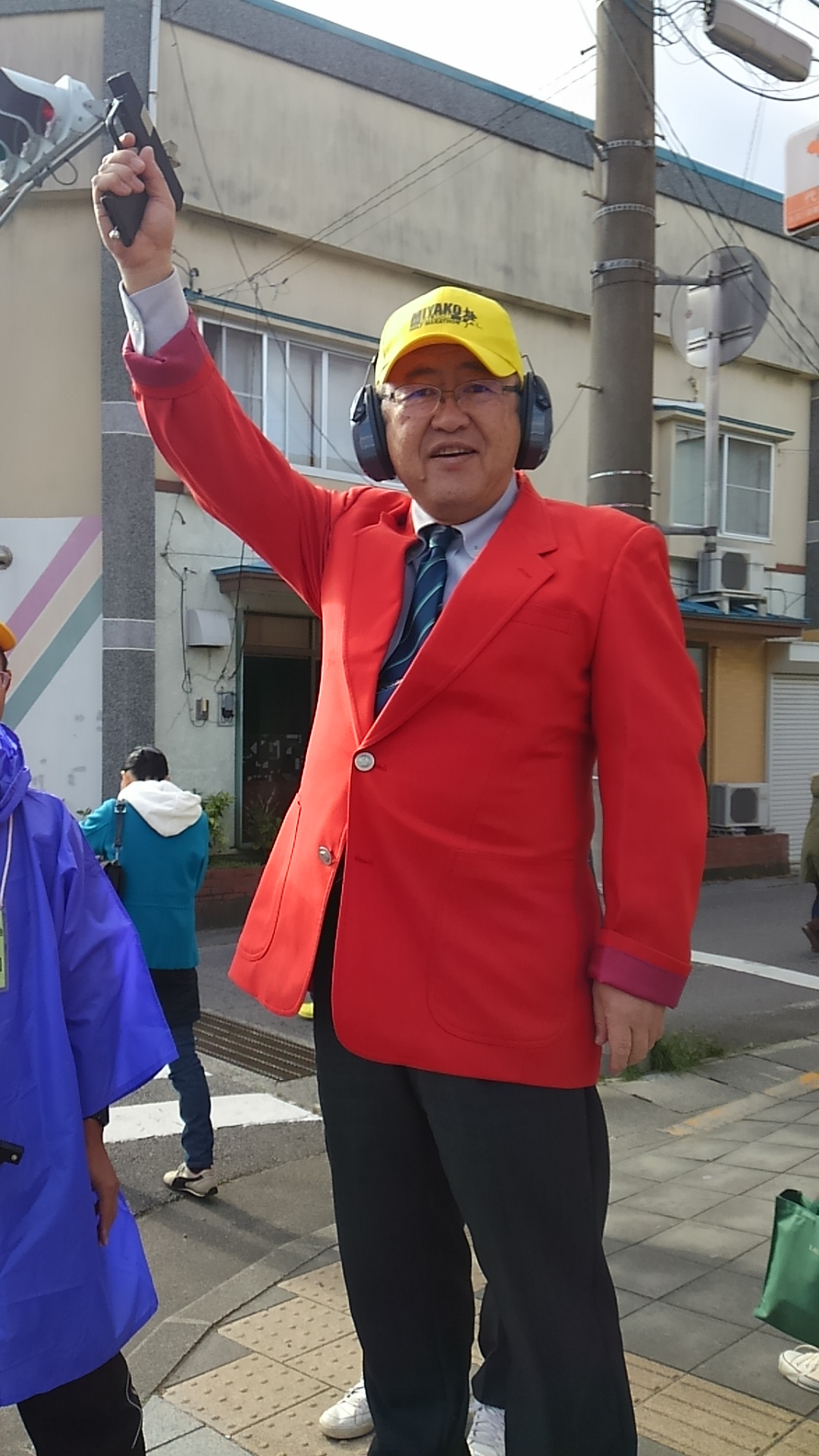 赤いジャケットを着て、黄色い帽子を被りヘッドホンを着けた市長が、右手に持ったピストルを上に掲げている写真