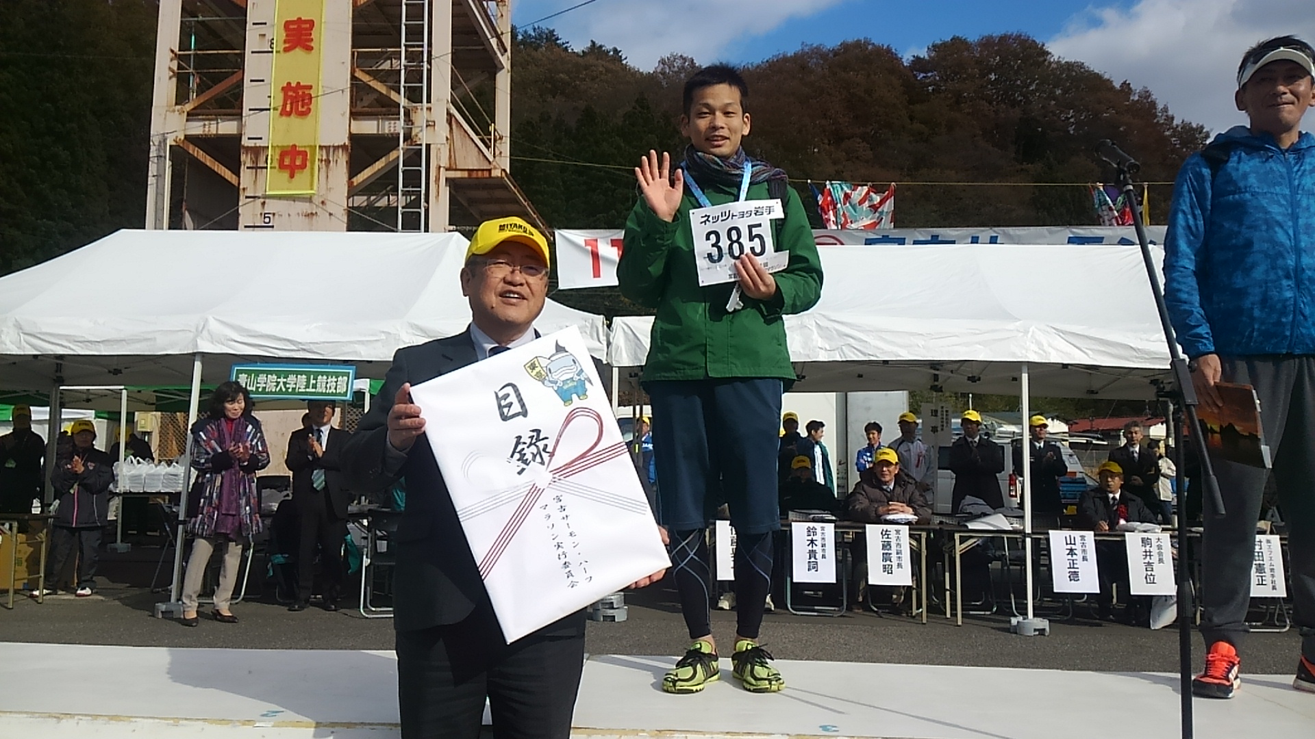 目録の大きいパネルを持つ市長の後方で、「385」と書かれたゼッケンをつけた男性が台の上に立っている写真
