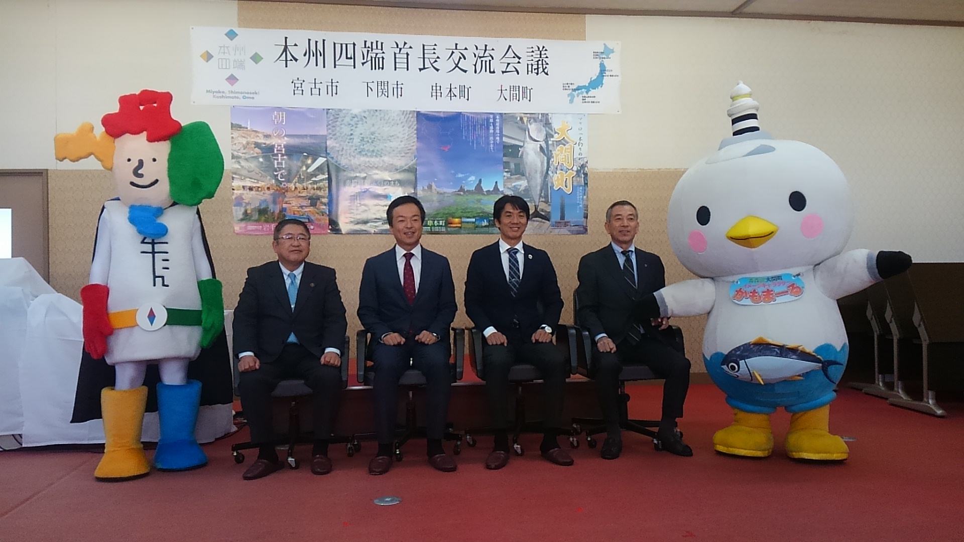 椅子に座った市長と3名の男性の両側によんたんとかもまーるが立って並ぶ記念写真