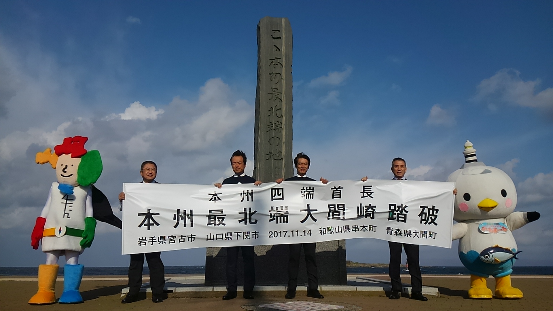 「本州四端首長 本州最北端大間崎踏破」と書かれた横断幕を持つ山本市長と3名の男性、両側に立つよんたんとかもまーるの記念写真