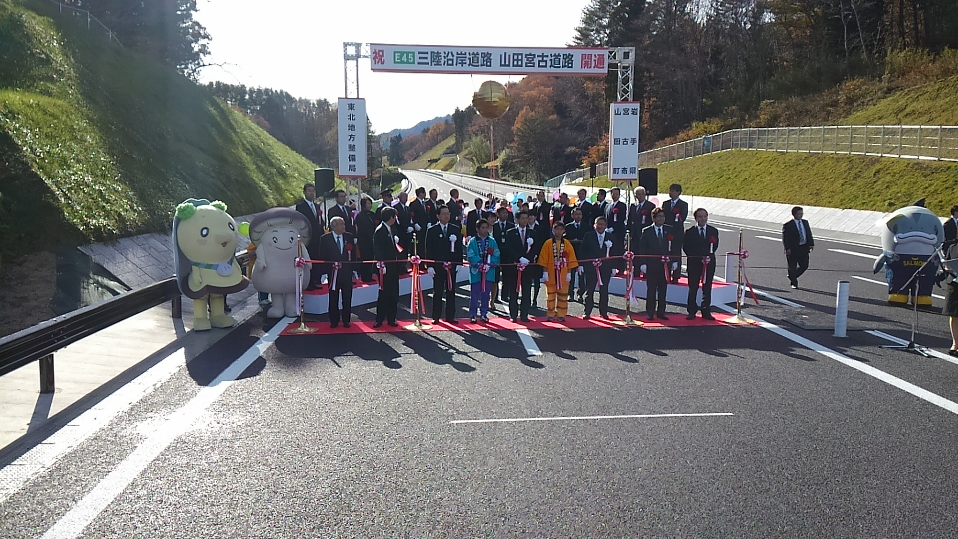 道路を横断するように敷かれたレッドカーペットの上でテープカットをする様子の方々の写真