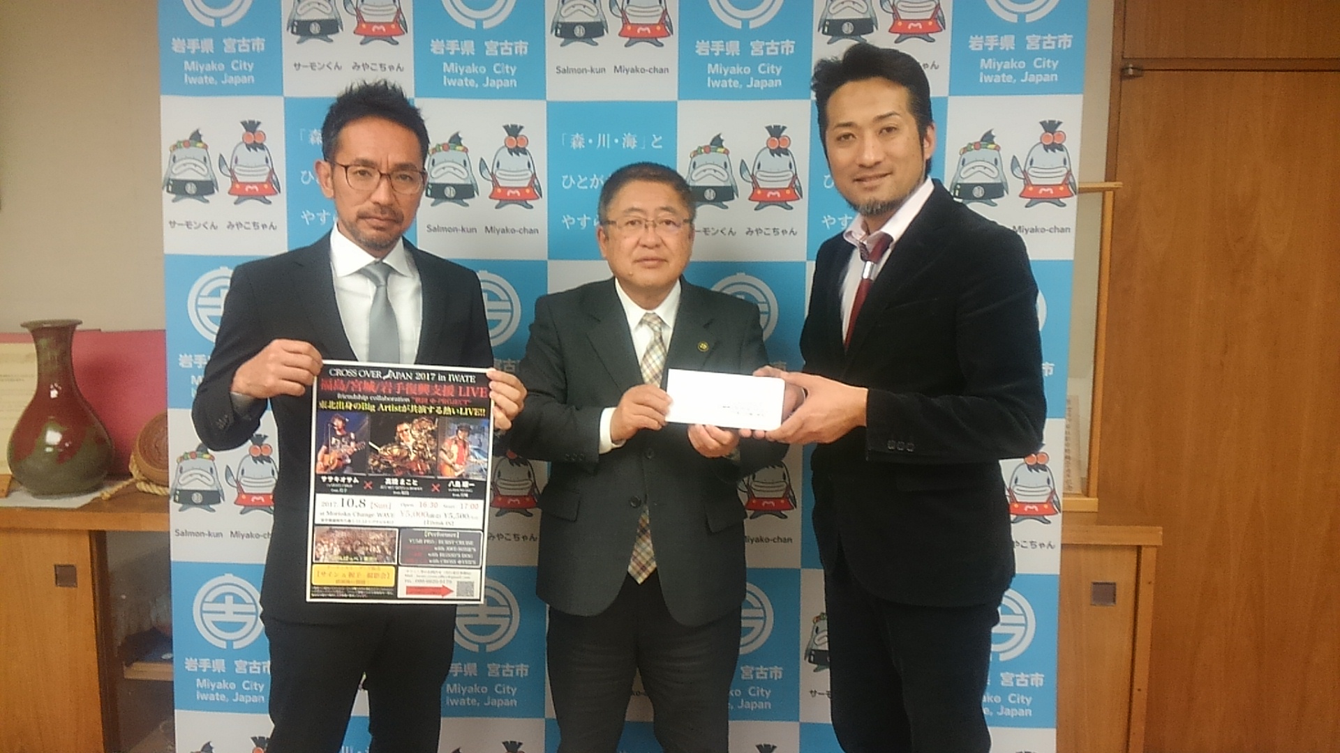バックパネルの前でポスターを持つ男性と、市長、男性1名が立って並ぶ記念写真