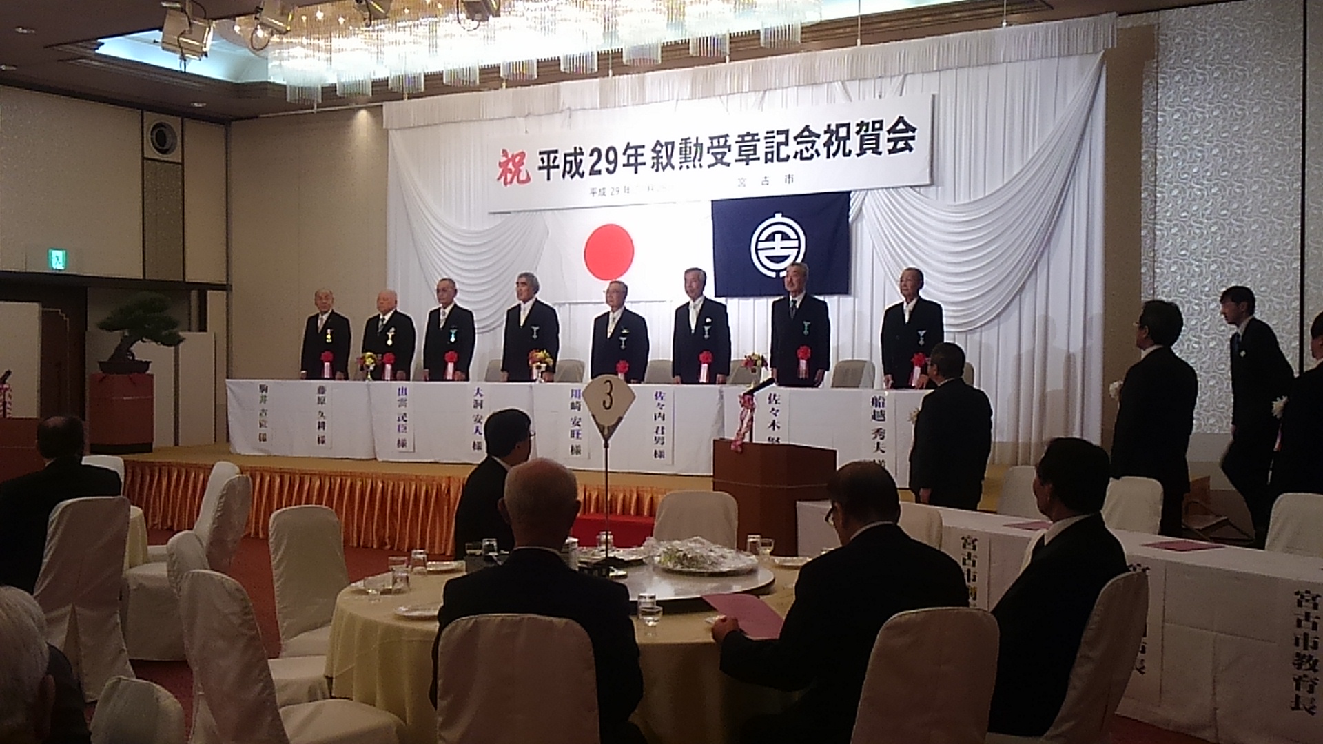 「祝 平成29年叙勲受章記念祝賀会」と書かれた横断幕、日本国旗、宮古市の市町村旗が掲げられた壁の前で、一列に並び立っている受賞者の方々の写真