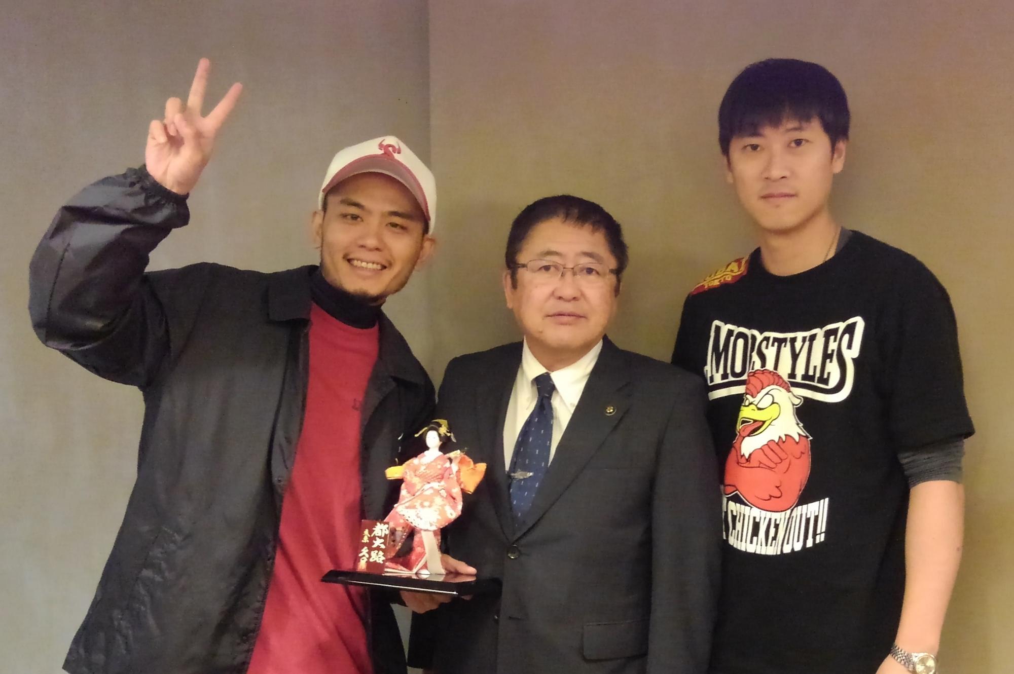 市長と右手でピースする男性、黒いTシャツを着た男性が並んで立つ記念写真