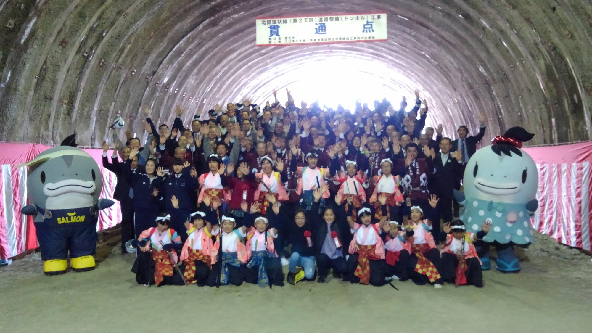 トンネル内で集合して写真を撮る大勢の関係者の方々と、両側に並ぶサーモンくんとみやこちゃんの写真