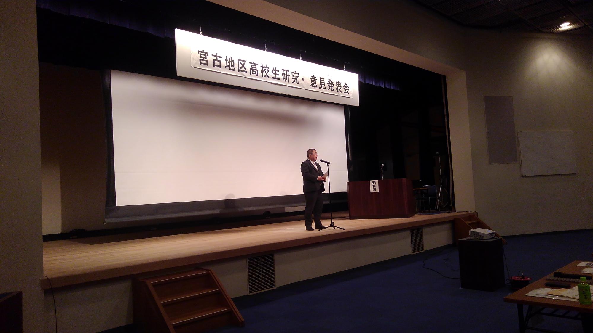 「宮古地区高校生研究・意見発表会」と書かれたパネルが吊るされた舞台上でスタンドマイクに向かい話をする市長の写真