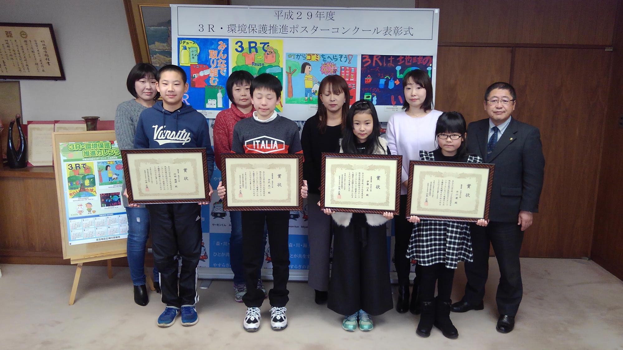 賞状の額縁を持つ4名の子供たちと、後方に並ぶ4名の女性と市長の写真