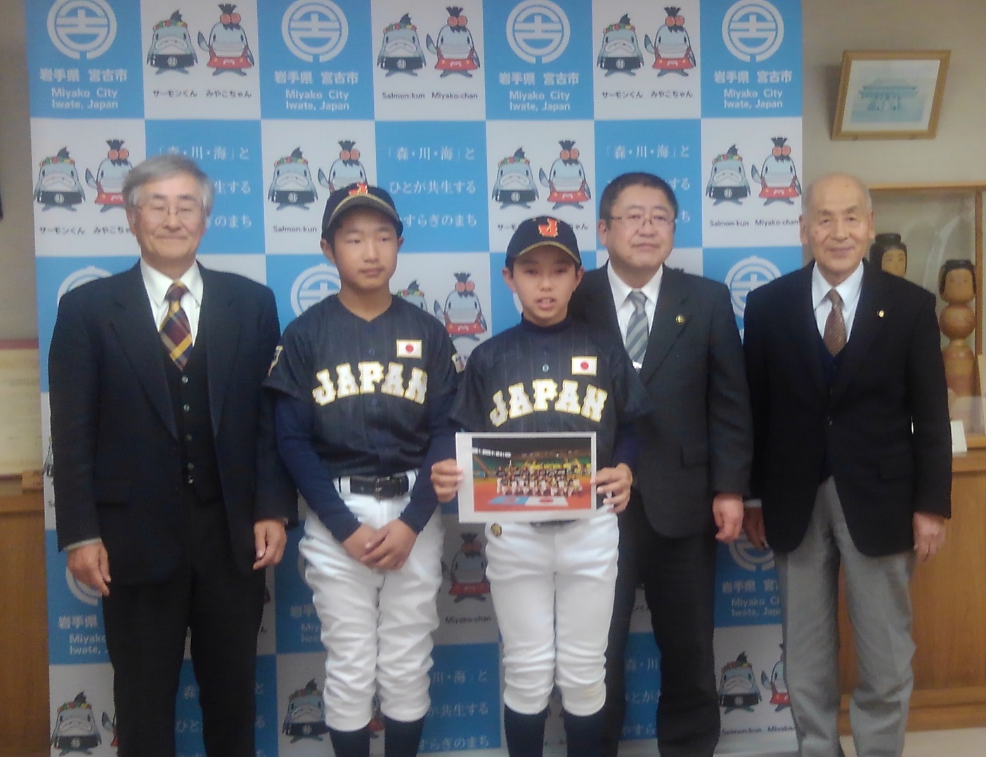 バックパネルの前で、野球のユニフォームを着た男の子2名、市長と関係者の男性2名の記念写真