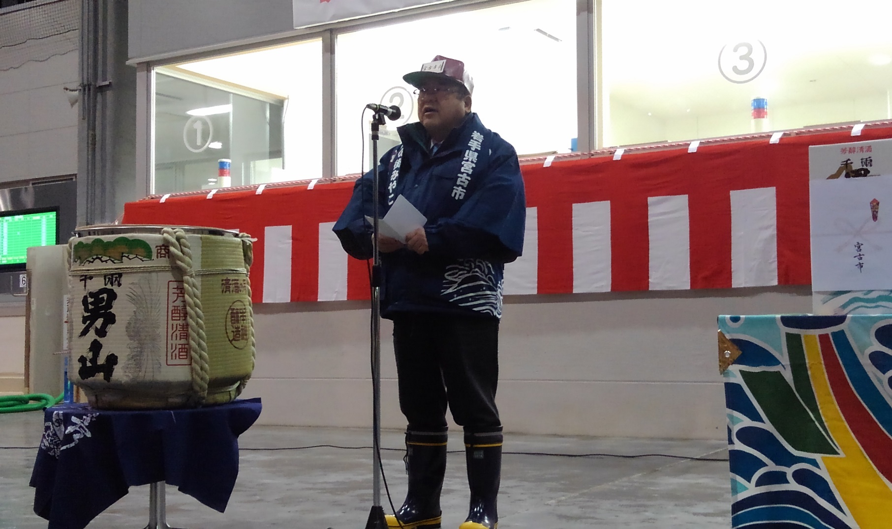 日本酒の大きな樽が置かれたテーブルの隣で、帽子を被り法被を着た市長がスタンドマイクに向かい話をする様子の写真