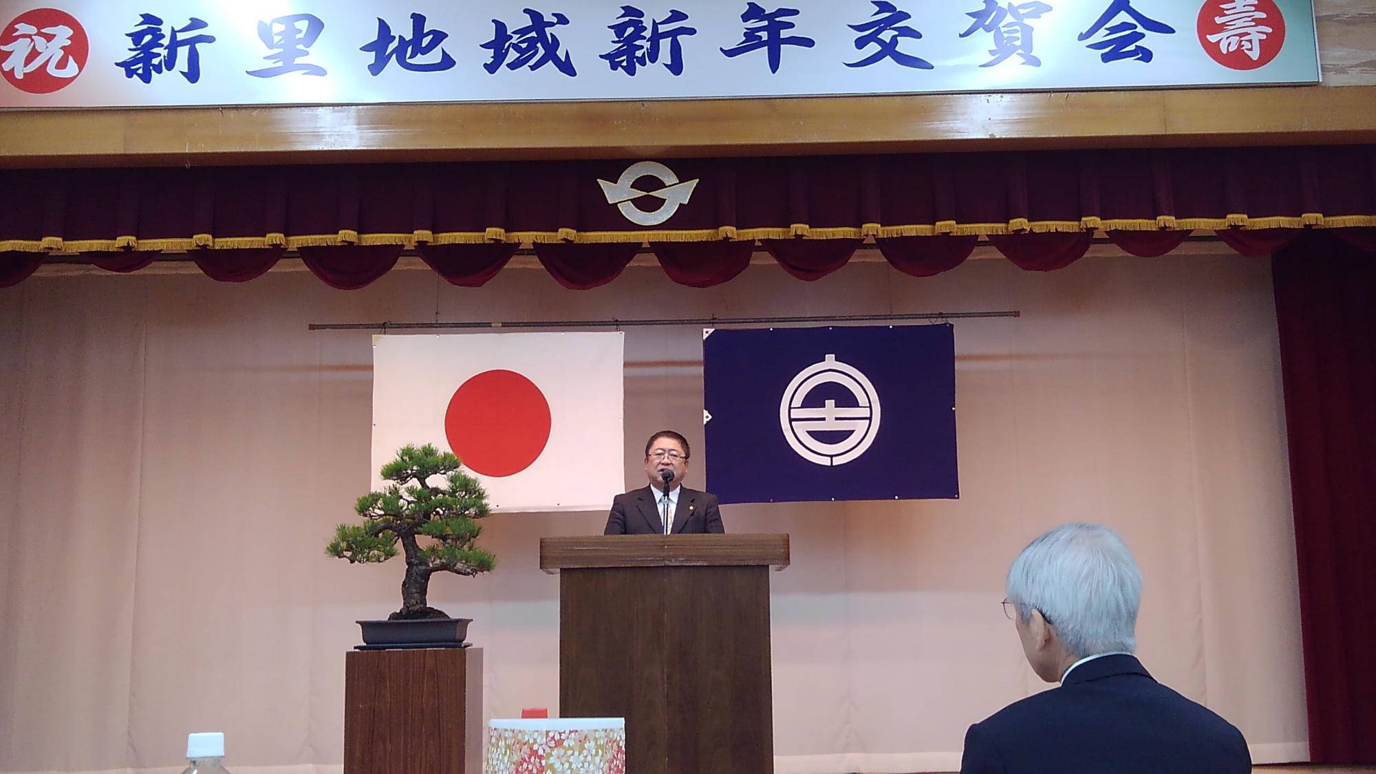 日本国旗と宮古市の市町村旗が掲げられ、盆栽が置かれた舞台で、演台に向かい話をする様子の市長の写真