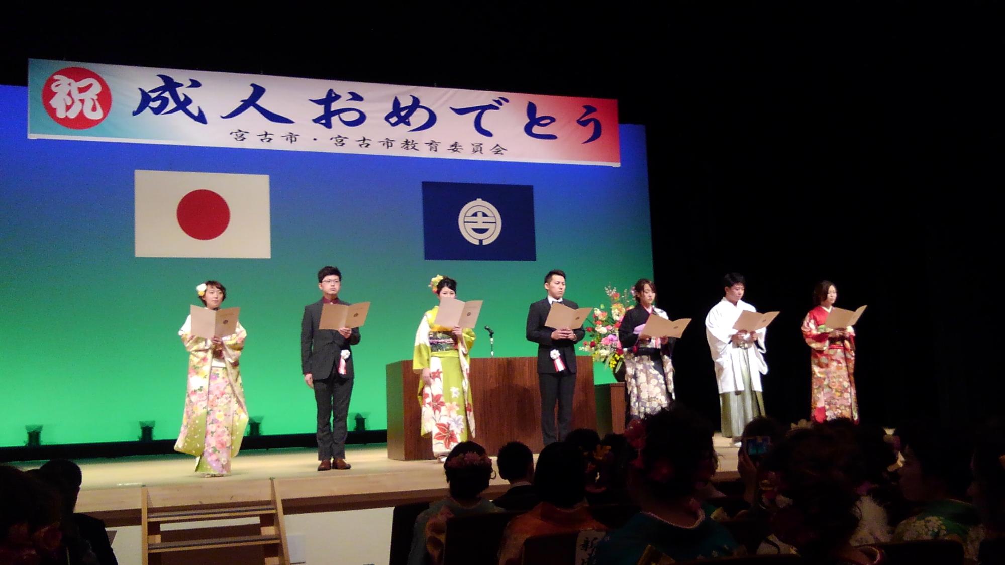 「祝成人おめでとう」と書かれた横断幕、日本国旗、宮古市の市町村旗が掲げられた舞台上で、着物を着た4名の女性、スーツを着た2名の男性、羽織袴を着た1名の男性が冊子を手に広げ横一列に並んでいる写真