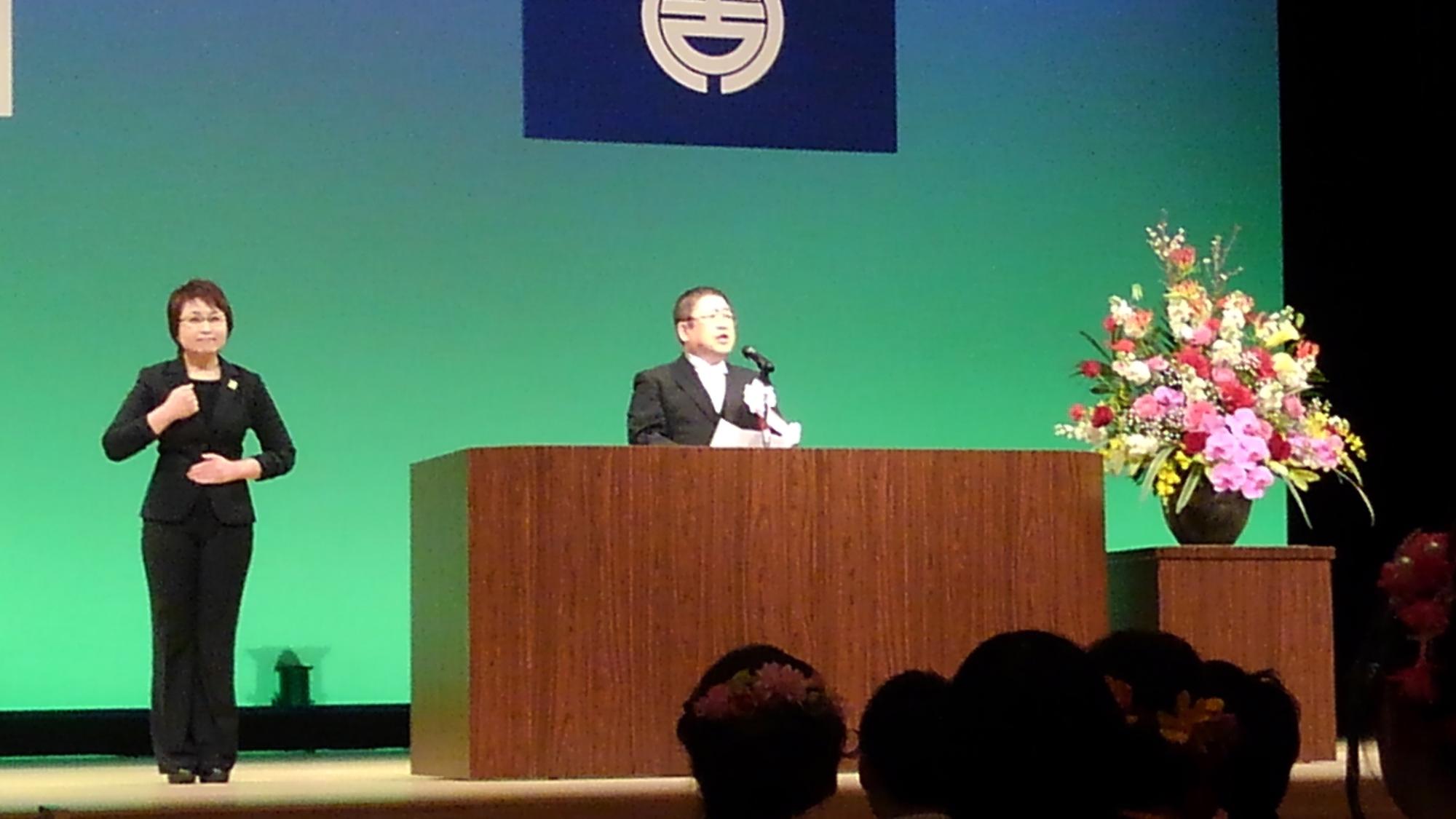 演台に置かれたマイクに向かい話をする様子の市長と、演台の横で手話通訳をしている様子の女性の写真