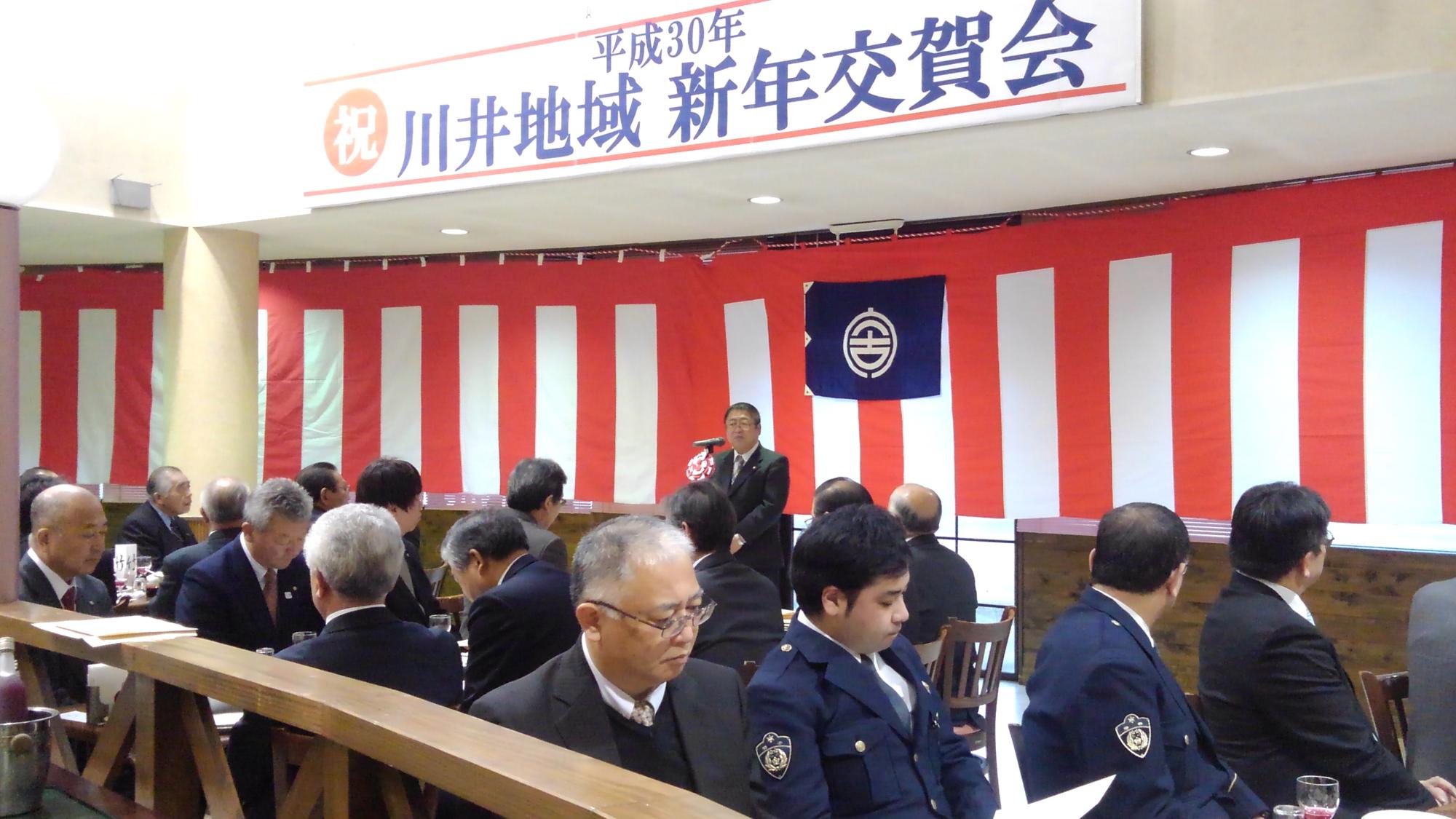 「祝川井地域 新年交賀会」と書かれた横断幕が吊るされた会場で、席につく関係者たちとスタンドマイクに向かい話をする様子の市長の写真