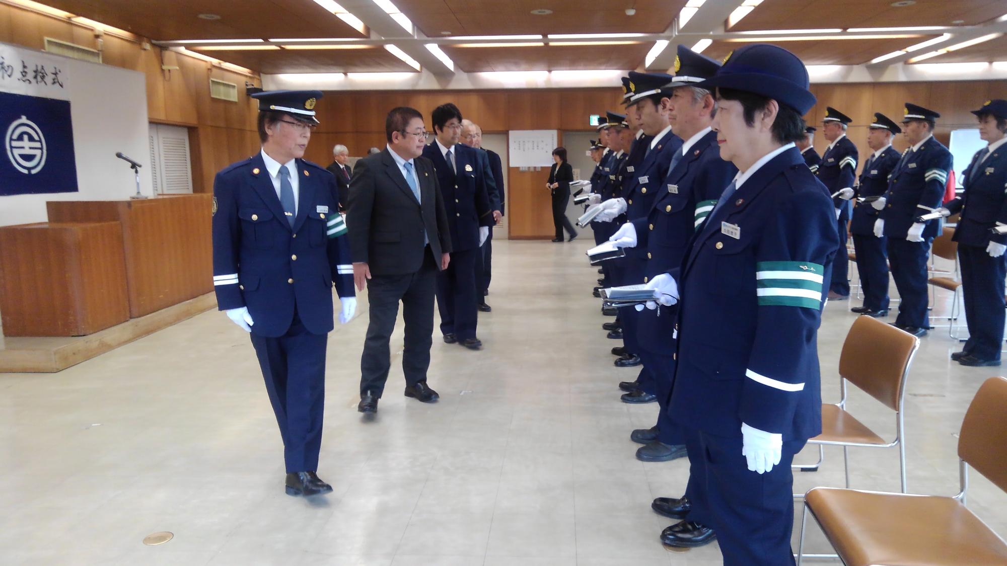 椅子の前に立って並ぶ制服を着た交通安全指導員の方々の前を歩く市長の写真