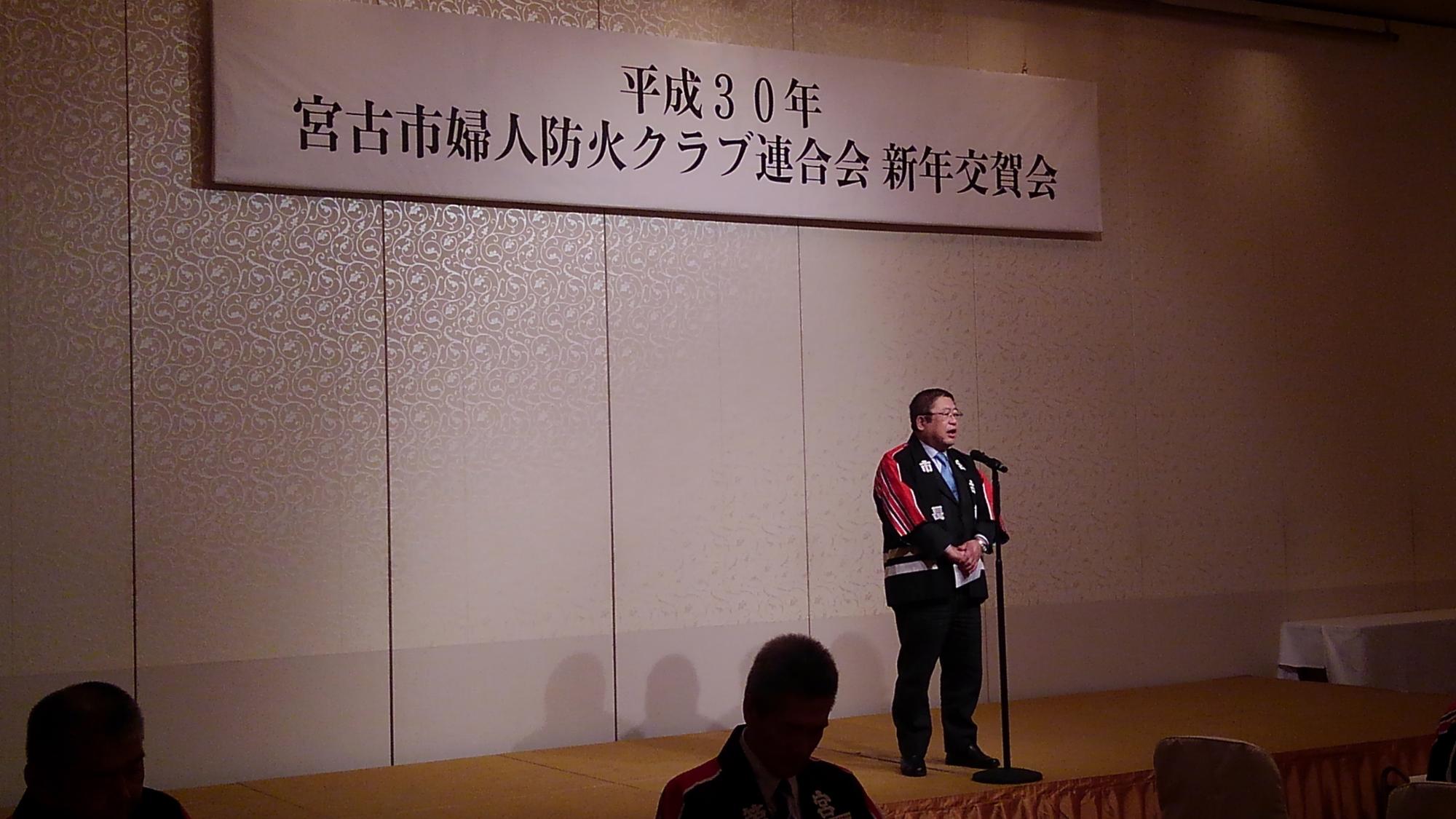 「平成30年 宮古市婦人科防火クラブ連合会 新年交賀会」と書かれた横断幕の下で、スタンドマイクに向かい話をする様子の市長の写真