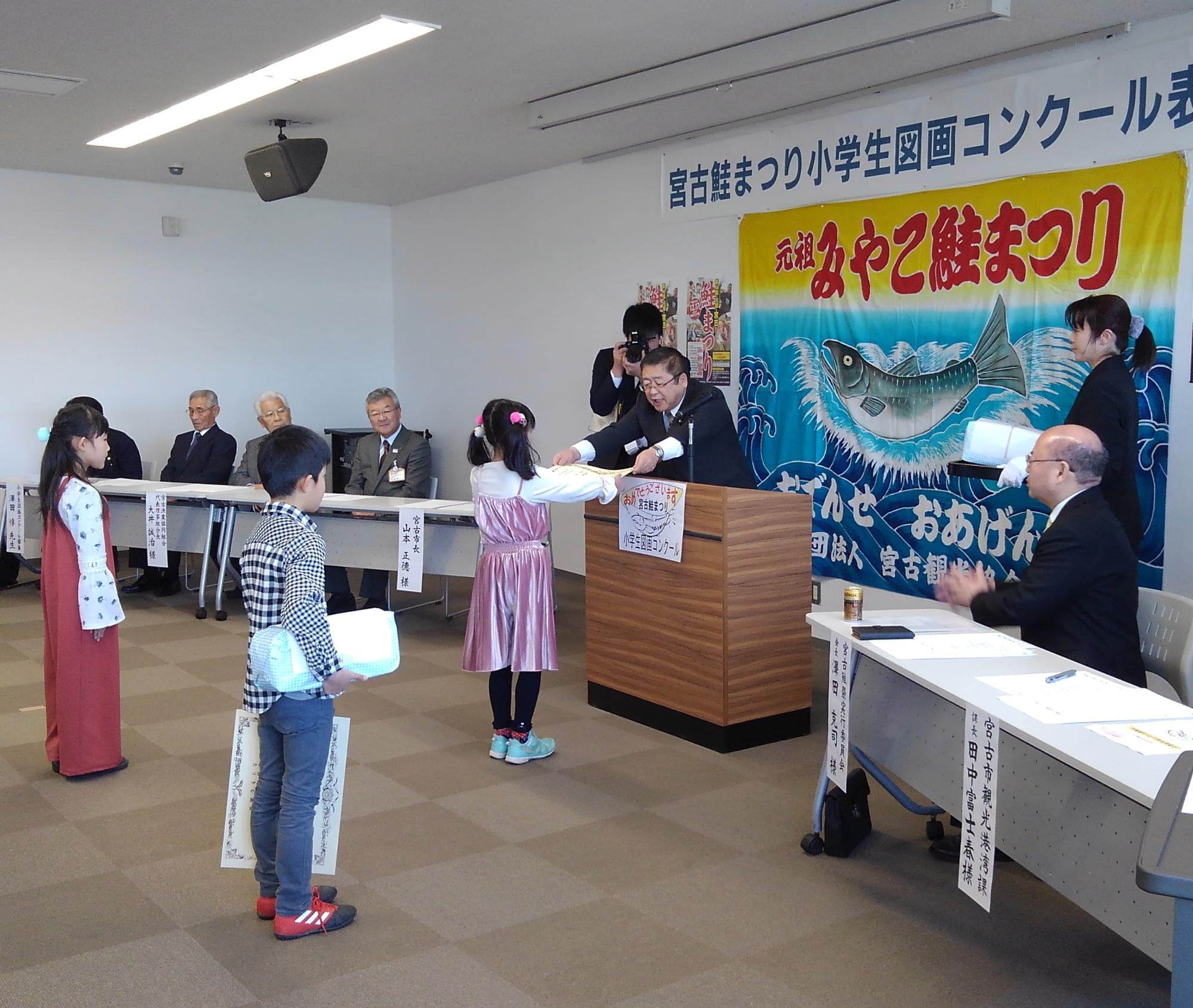 演台越しに賞状を渡す市長と、両手で受け取るピンクのワンピースを着た小学生の写真