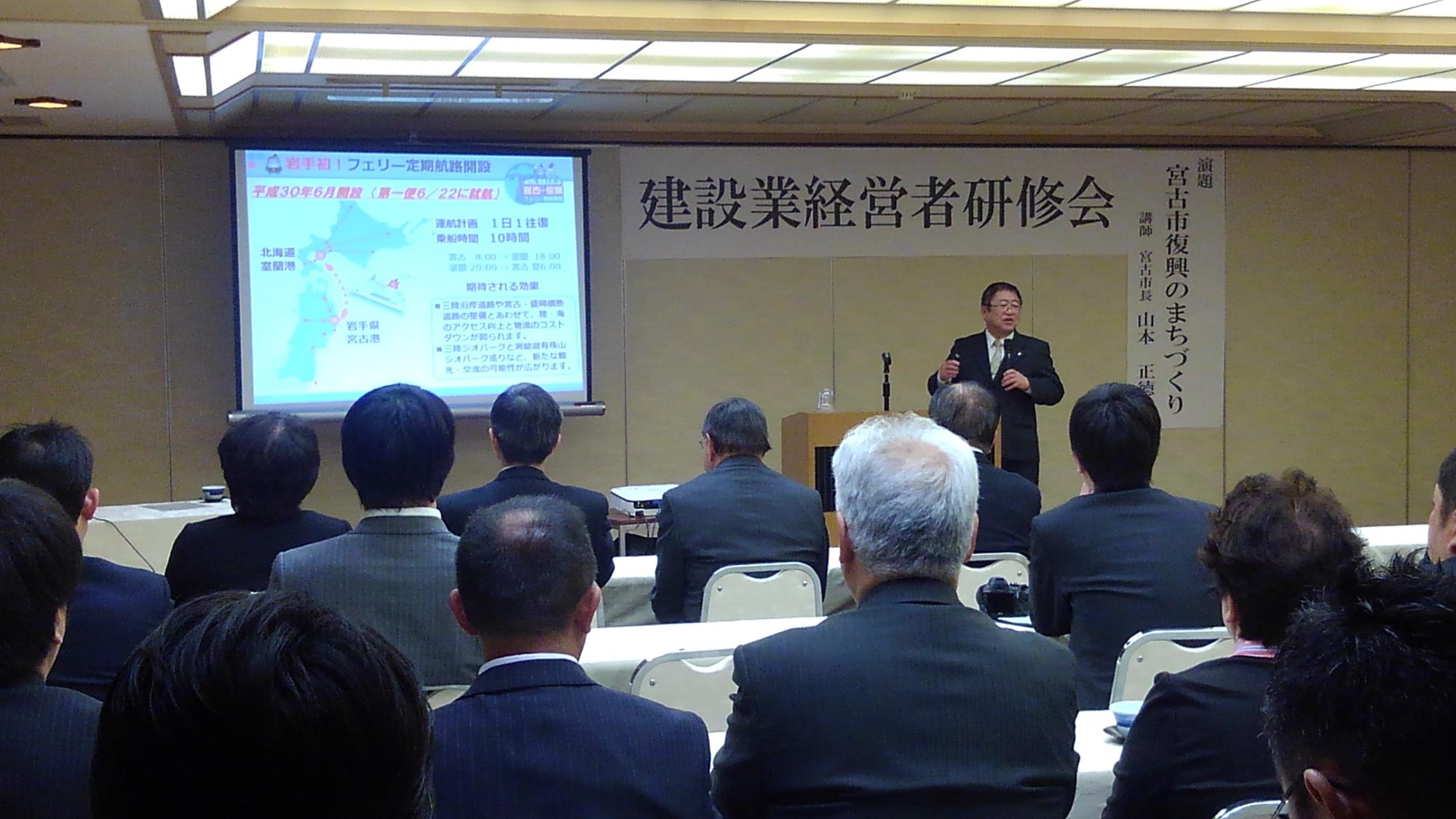 画面が映されたスクリーンの横で、左手にマイクを持ち話をする様子の市長と、並べられたテーブルの席に着く関係者の方々の写真
