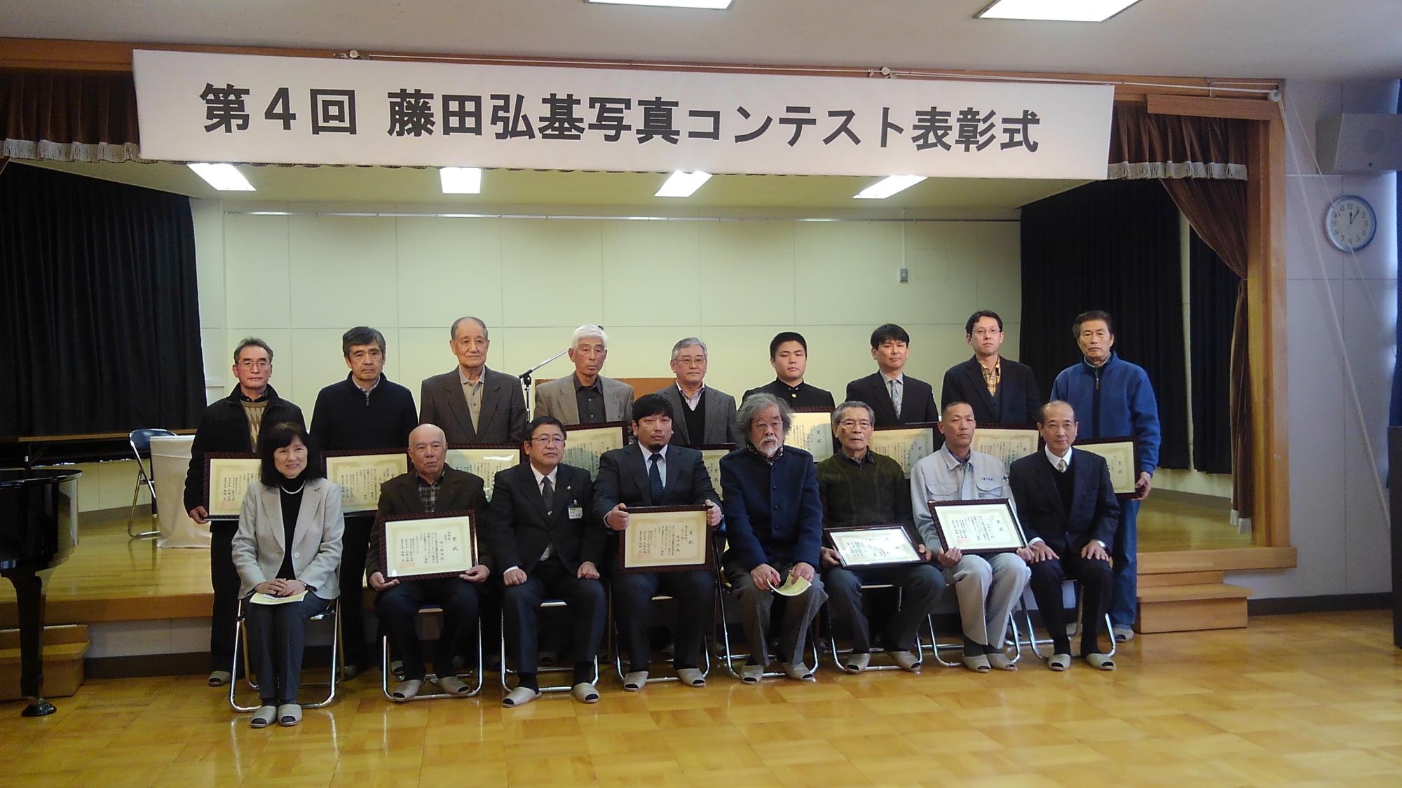 「第4回 藤田弘基写真コンテスト表彰式」と書かれた横断幕の下で、1列目に椅子に座った8名、2列目で立つ9名が並んで写る記念写真