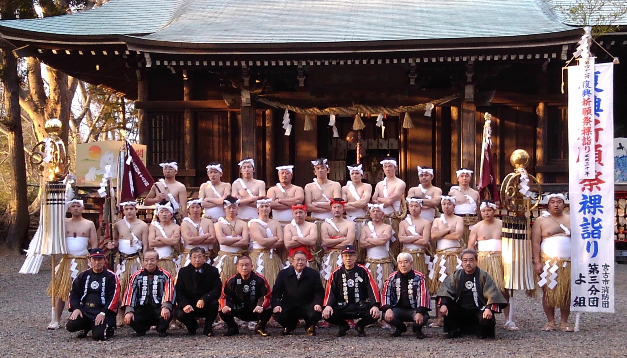 神社の前で上半身裸で鉢巻と腰蓑を巻き両腕を胸の前で組む男性たちと、法被を来た男性たち、スーツを着た市長が3列になって並んでいる記念写真