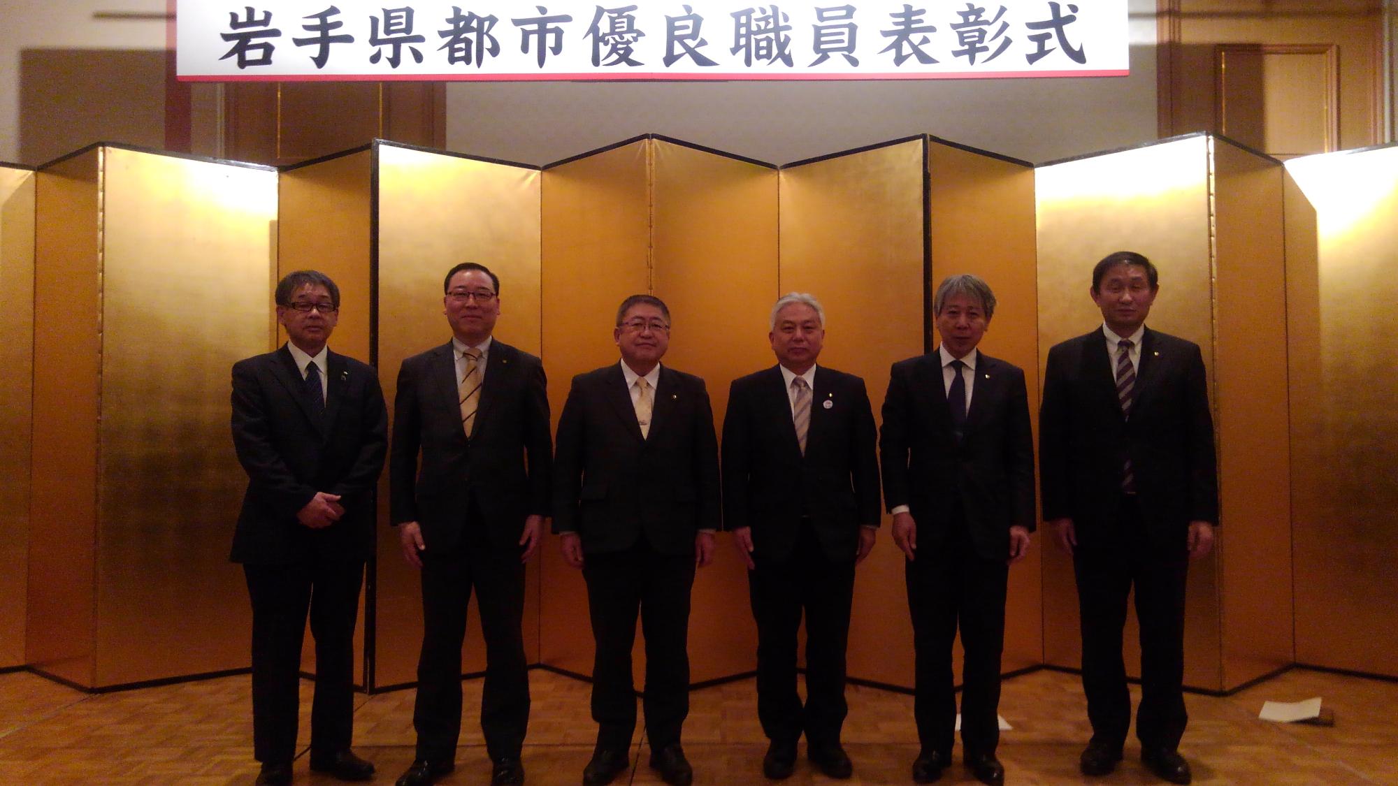 「岩手県都市優良職員表彰式」と書かれた横断幕の下で、金屏風の前に横一列に並ぶ市長と5名の男性の写真