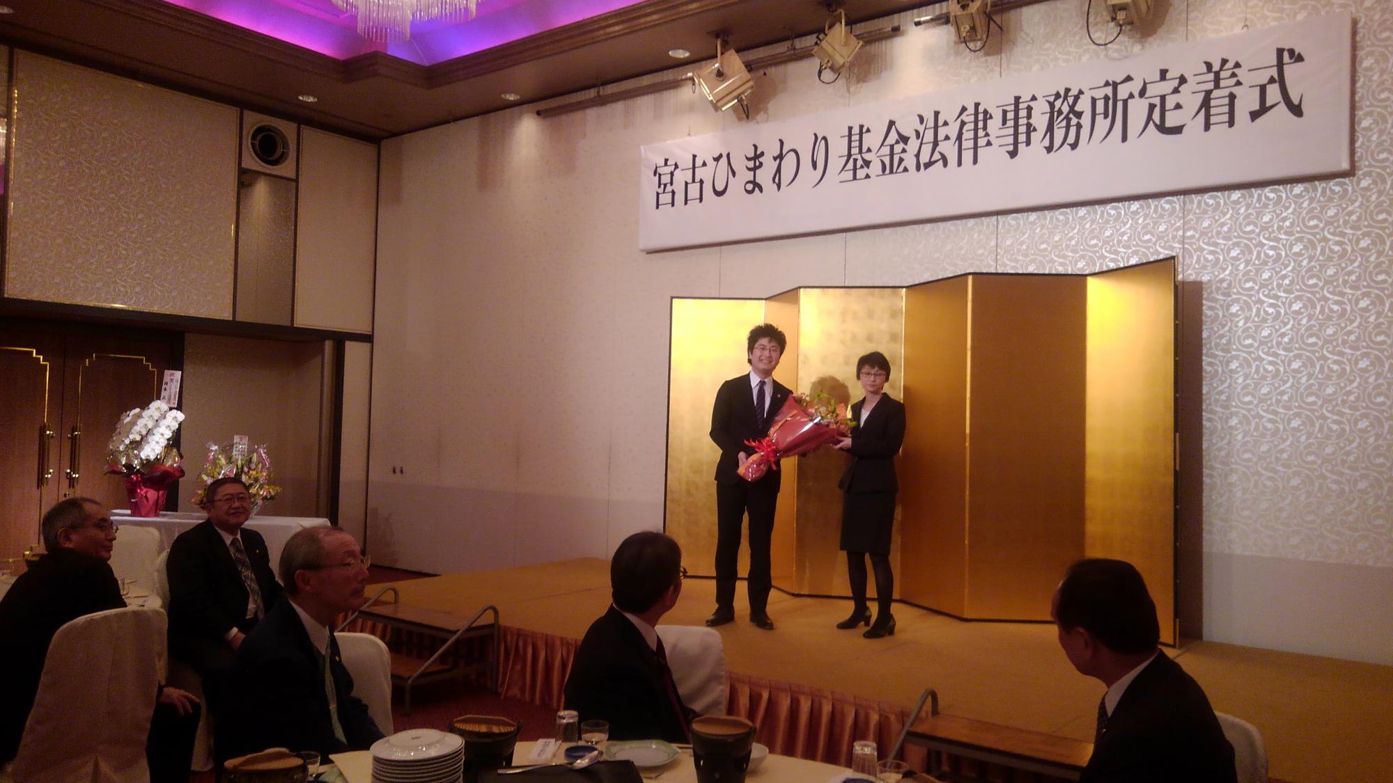 「宮古ひまわり基金法律事務所定着式」と書かれたパネルの下にある金屏風の前で、花束を持つ男性と女性が立っている写真