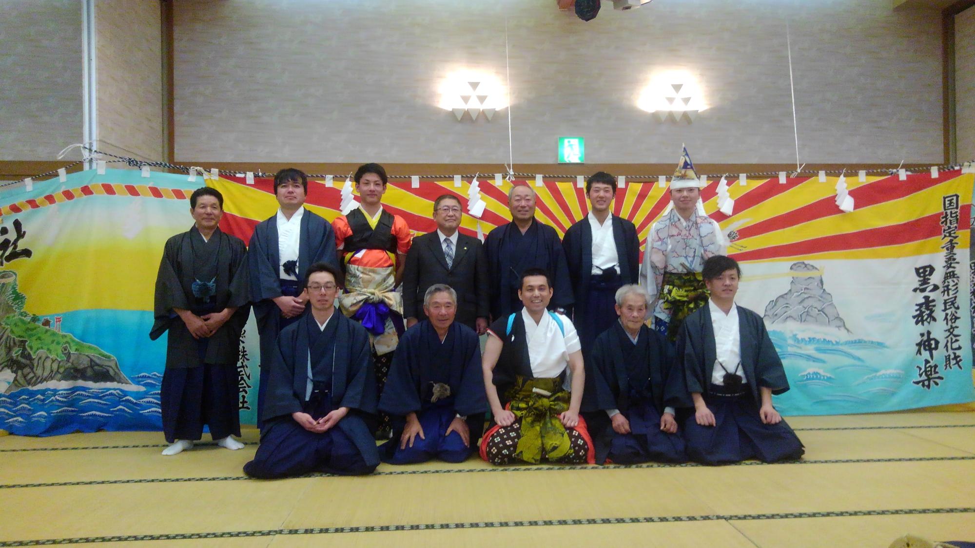 畳の上で、前列で正座をする5名と2列目で直立する市長と6名の関係者の男性たちの写真