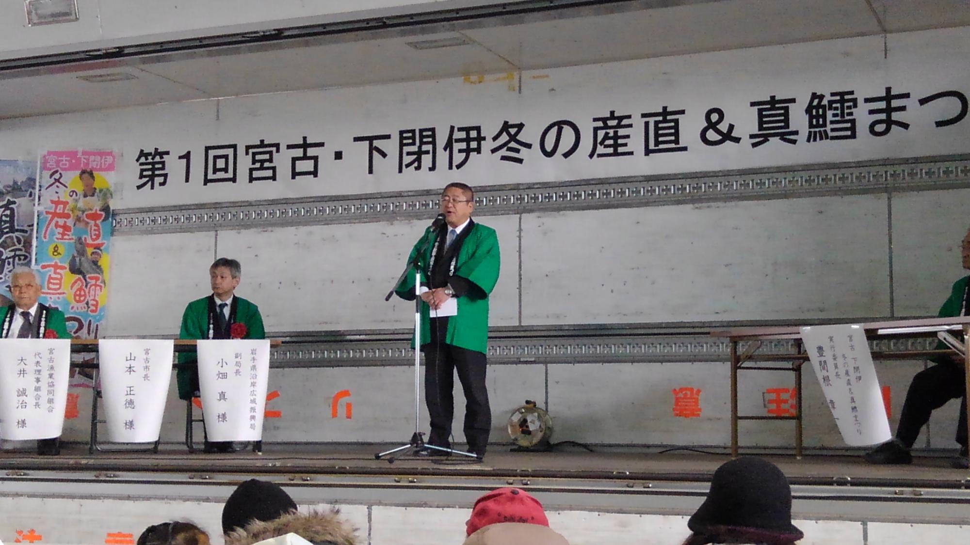 緑色の法被を着た市長がスタンドマイクに向かい話をしている様子の写真