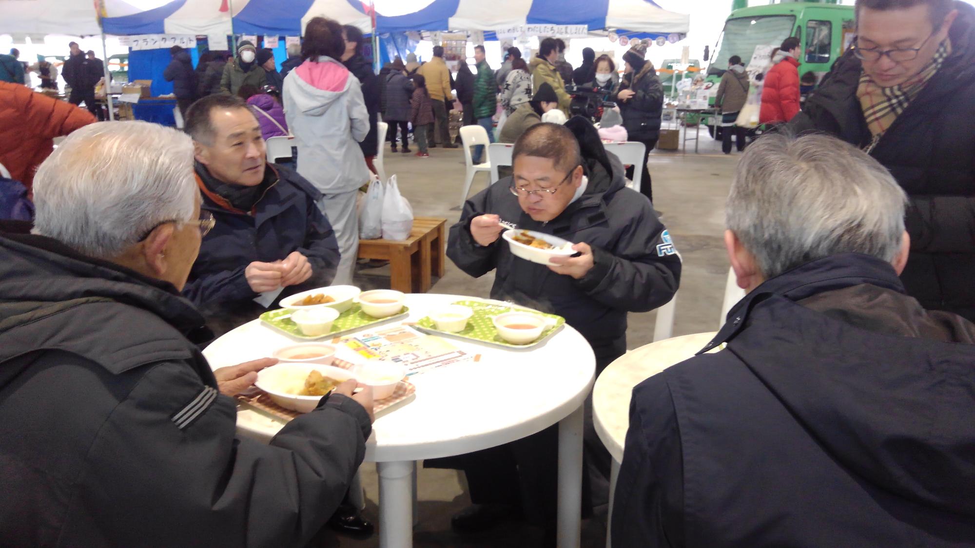 丸いテーブルに置かれた料理を食べている様子の市長と関係者の男性たちの写真