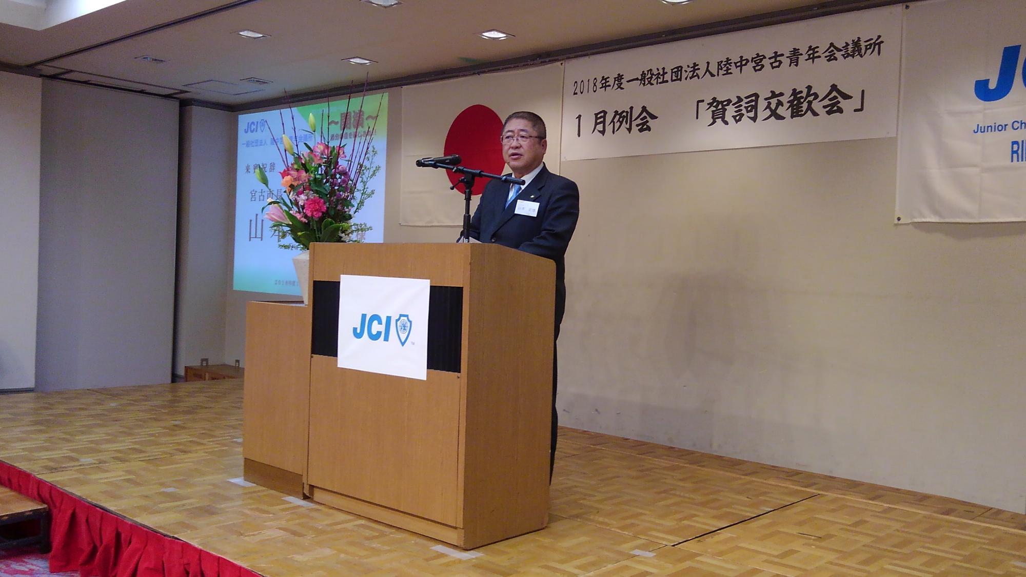 舞台に置かれた演台のマイクに向かい話をする様子の市長の写真