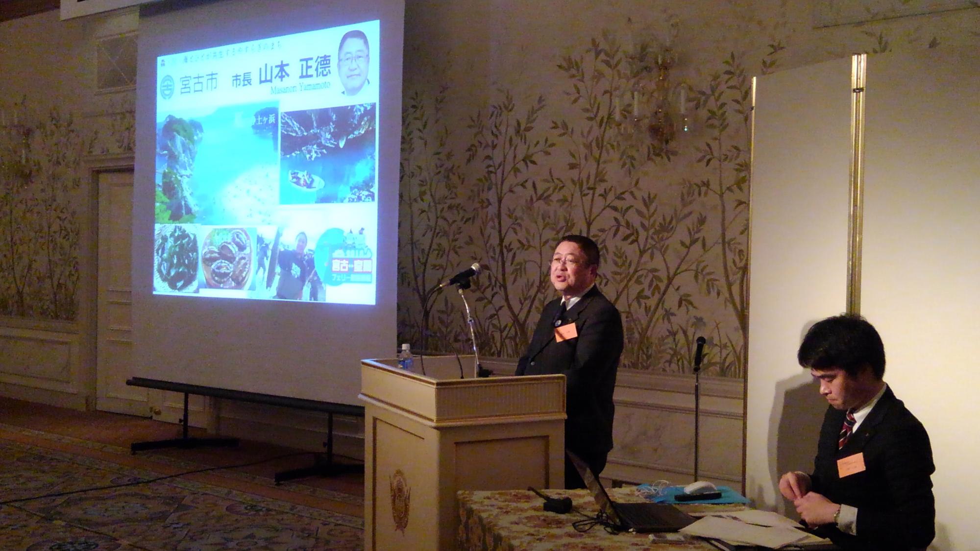 画面が映されたスクリーンの横の演台で話をする様子の市長の写真